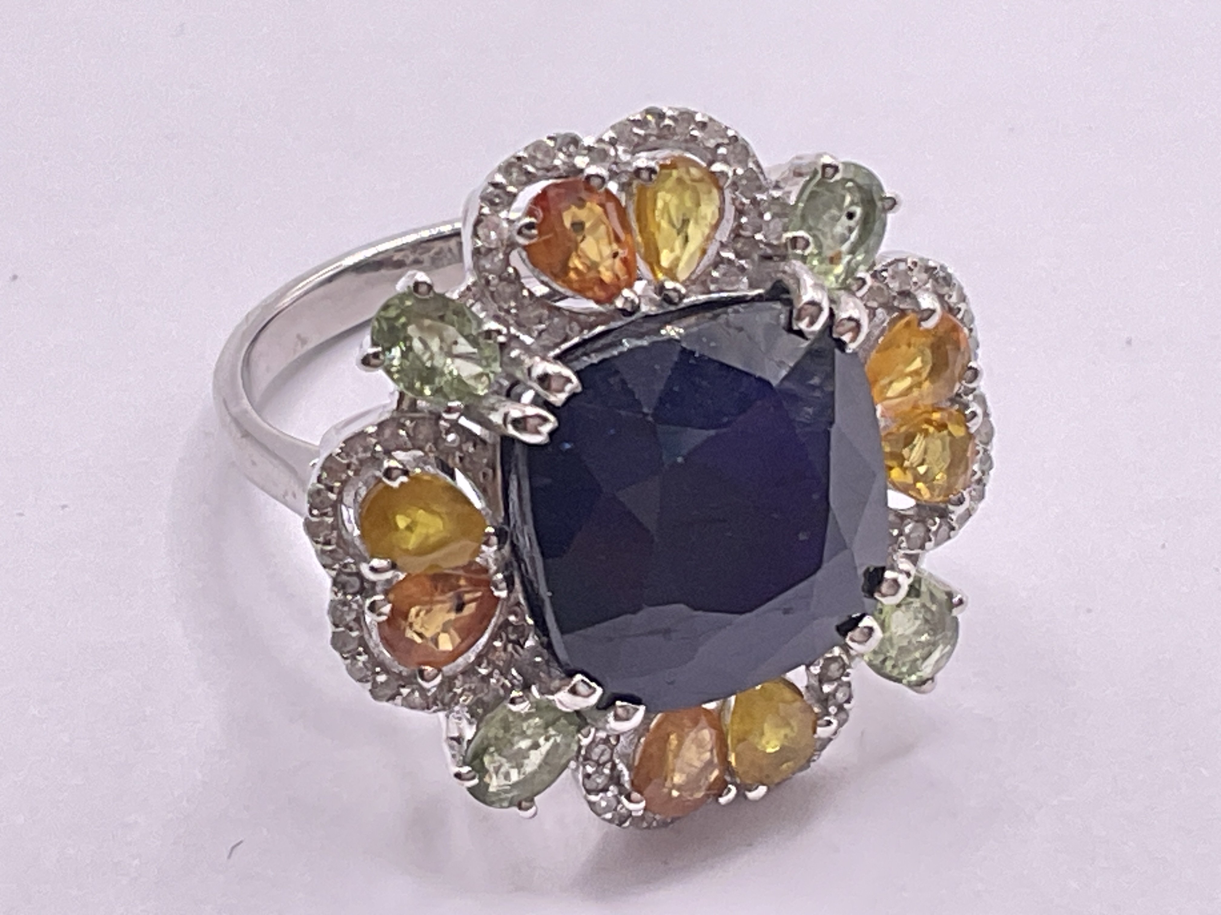 Floral style silver dress ring set with a central cushion cut blue sapphire (glass filled), surrounded by multi coloured sapphires and diamonds. Sapphires 12.00ct total, approx. Diamonds 0.40ct approx. Ring size approx N. Postage category A.