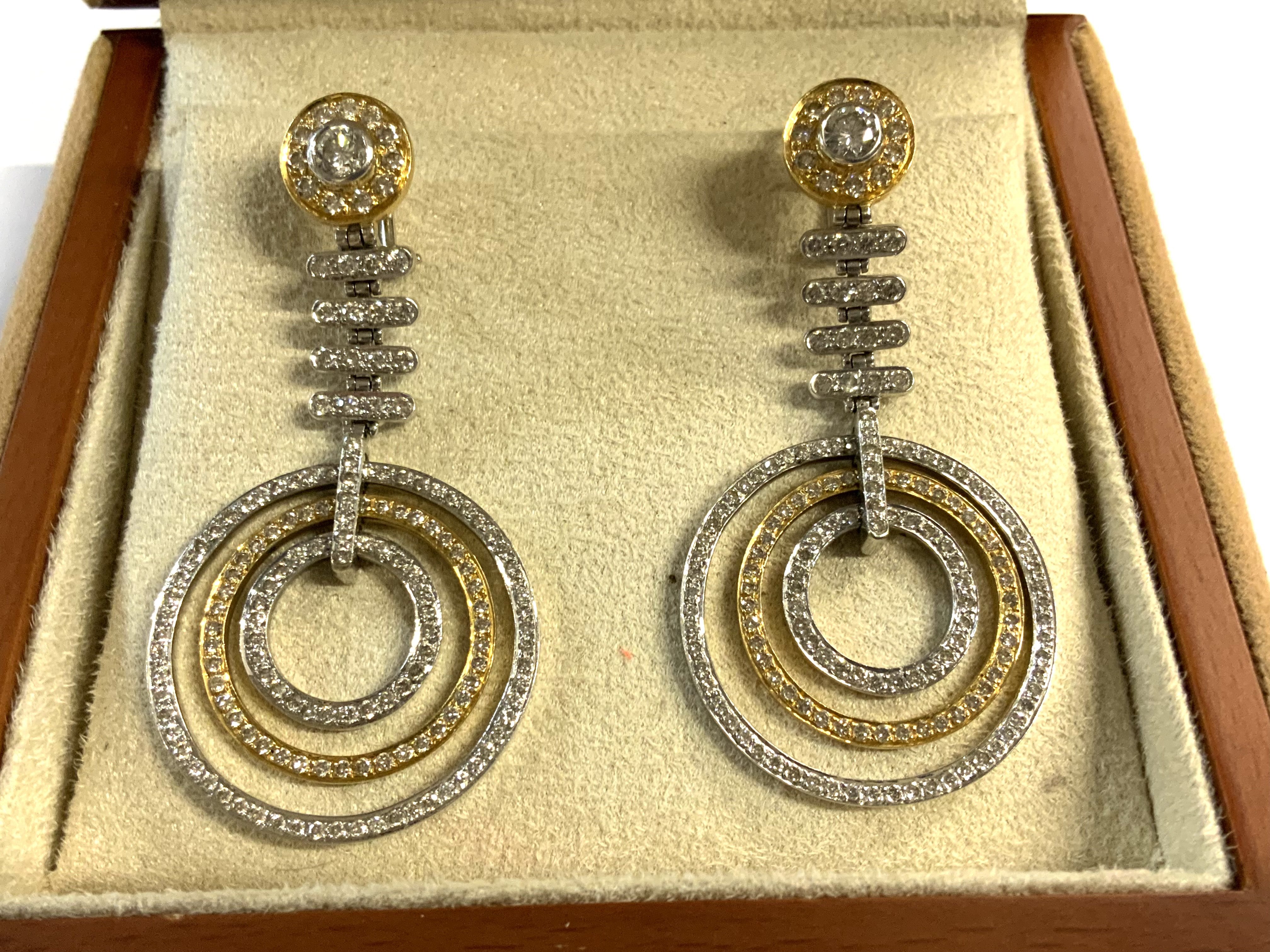 A pair of 18ct yellow and white gold drop earrings each stud section set with a 0.33ct solitare diamond surrounded by small 0.01ct stones, the drop section in white gold also inset with cut stones finished by 3 concentric rings each encrusted with small cut stones. Purchased at Gomez & Molina, Joyeros, Spain. (A)