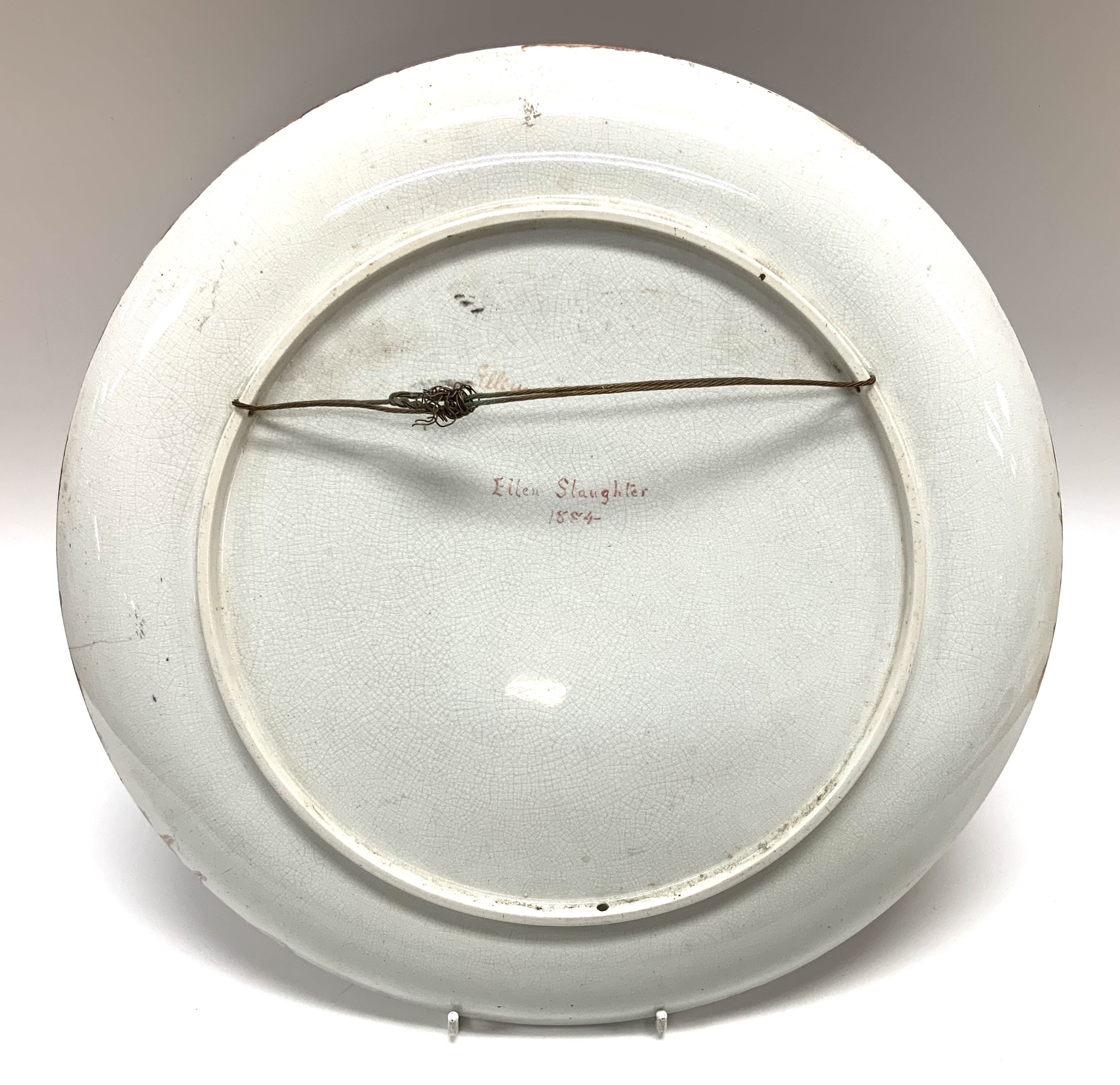 A large earthenware pottery charger with hand painted view of a lady using peacock feathers. Named and dated to reverse 1884 Ellen Slaughter. 36.5cm (D)