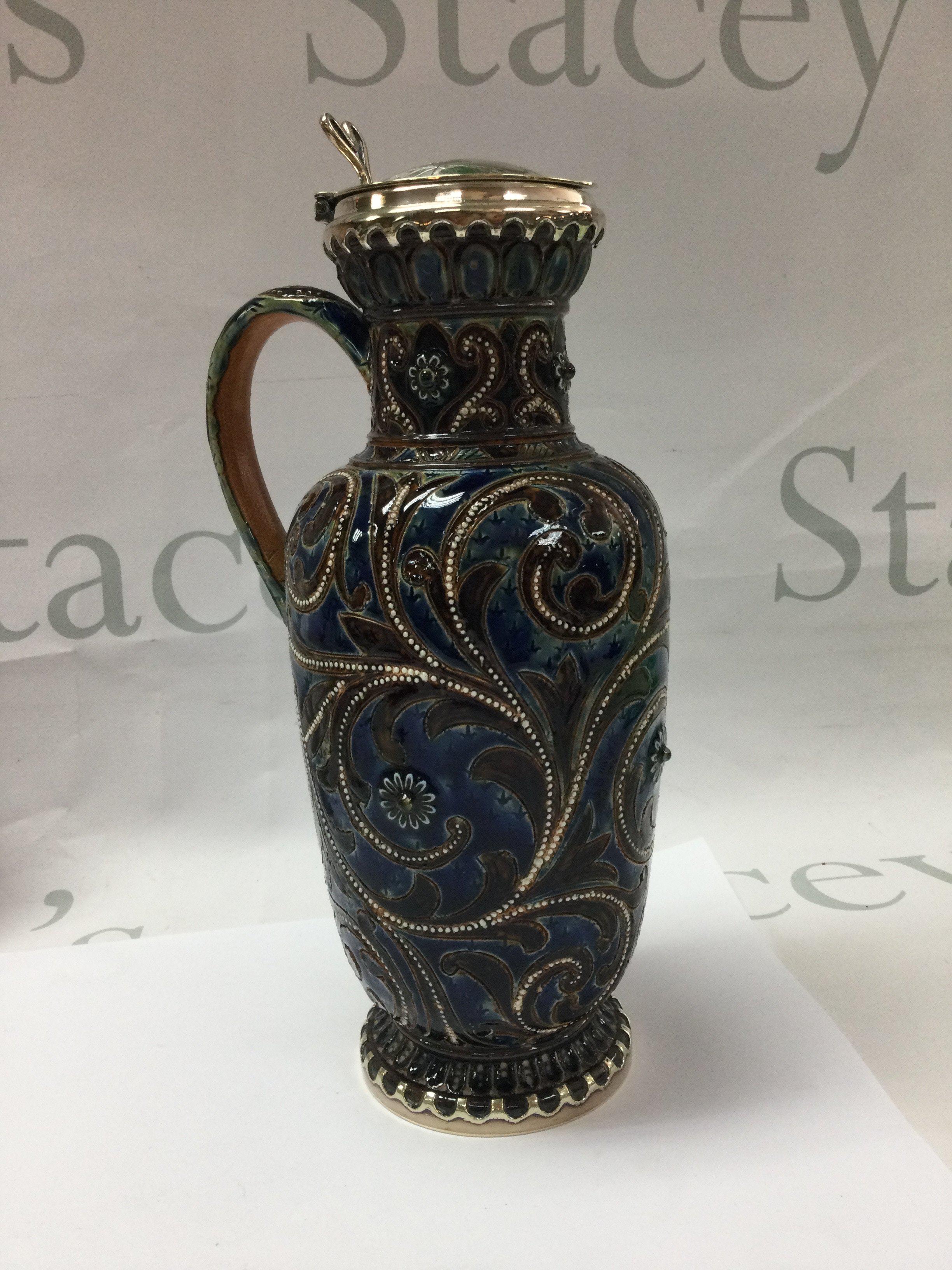 A Doulton Lambeth stoneware jug decorated with flower heads with silver plated lid 22 cm .