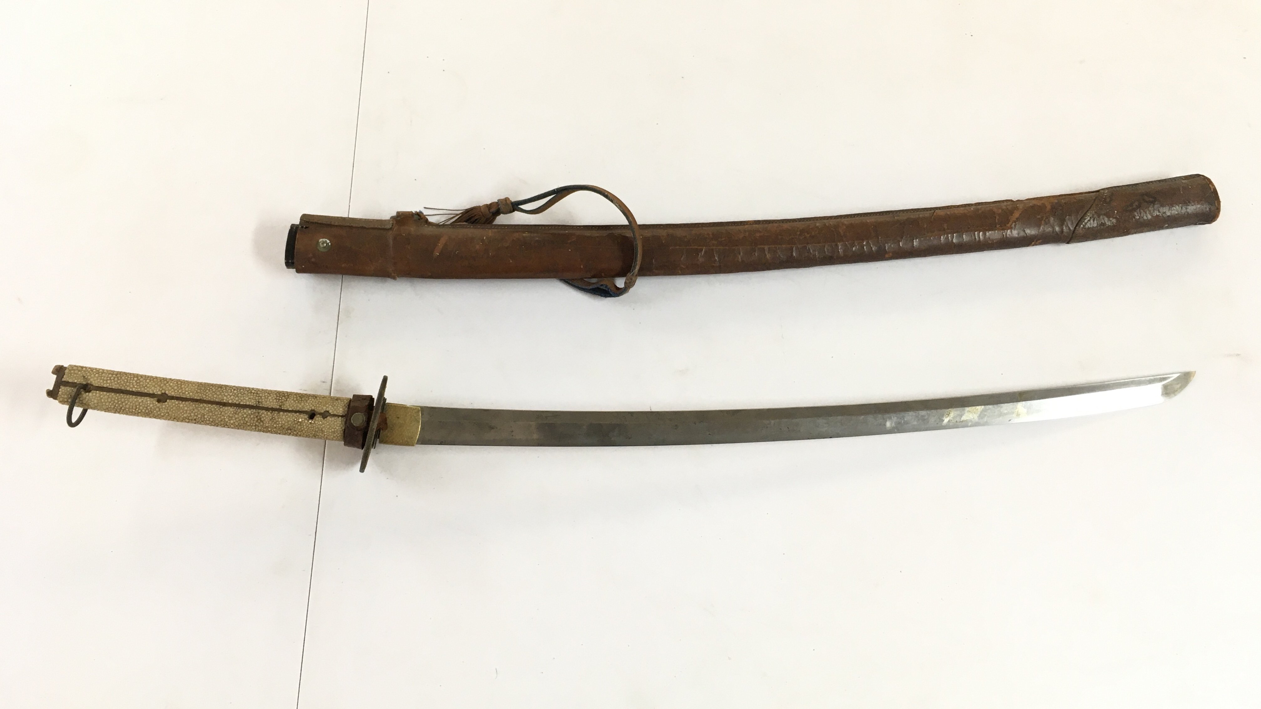 A WW2 Issue Japanese Katana with a ray skin wrapped handle and a leather scabbard, in need of restoration. Pin missing. Blade 66cm long, handle approximately 24cm long. Total length 90cm