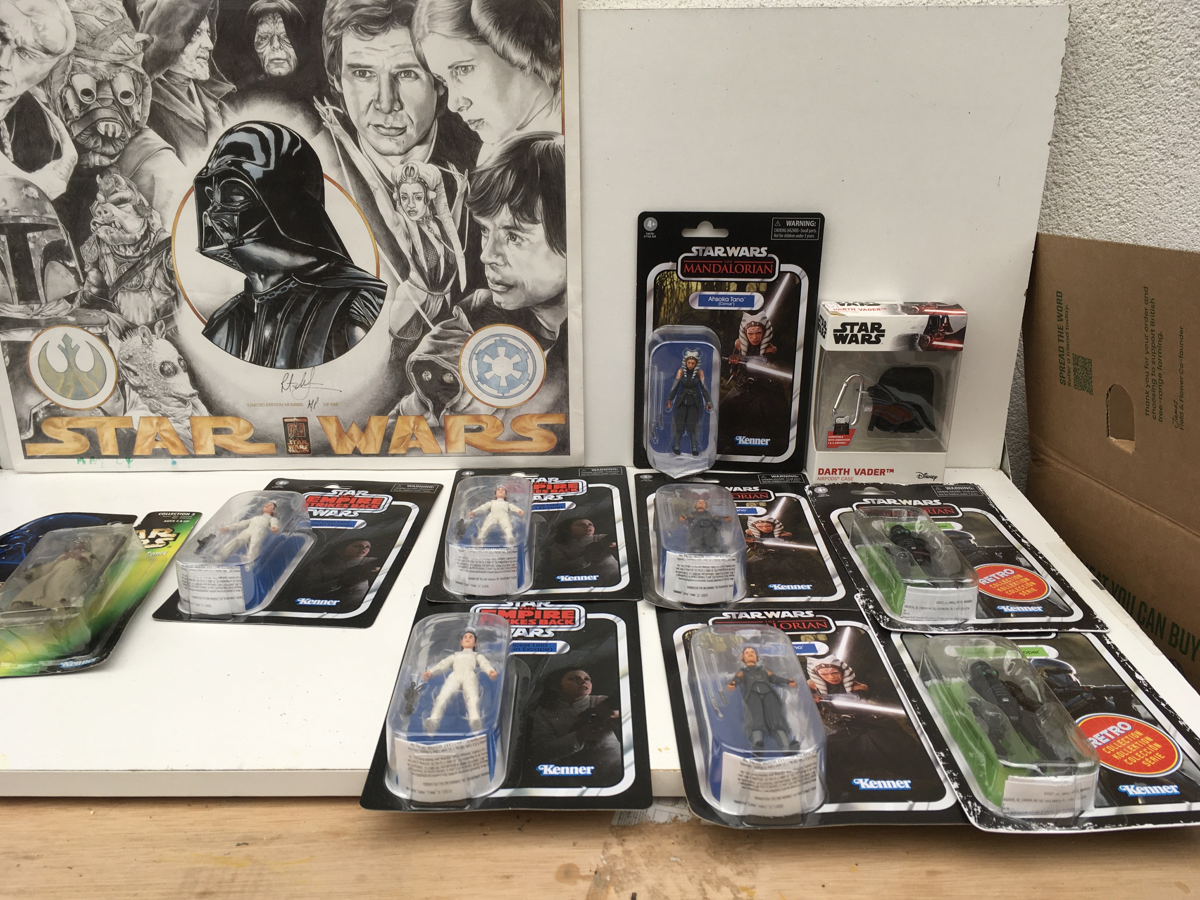 A group of 11 Star Wars pieces featuring a signed Artist Poof print from a limited edition of 1000 with Certificate of Authenticity . 9 x carded figures featuring the Empire Strikes Back..the Mandalorian and the Power of the Force. Also boxed Darth Vader AirPods Case.