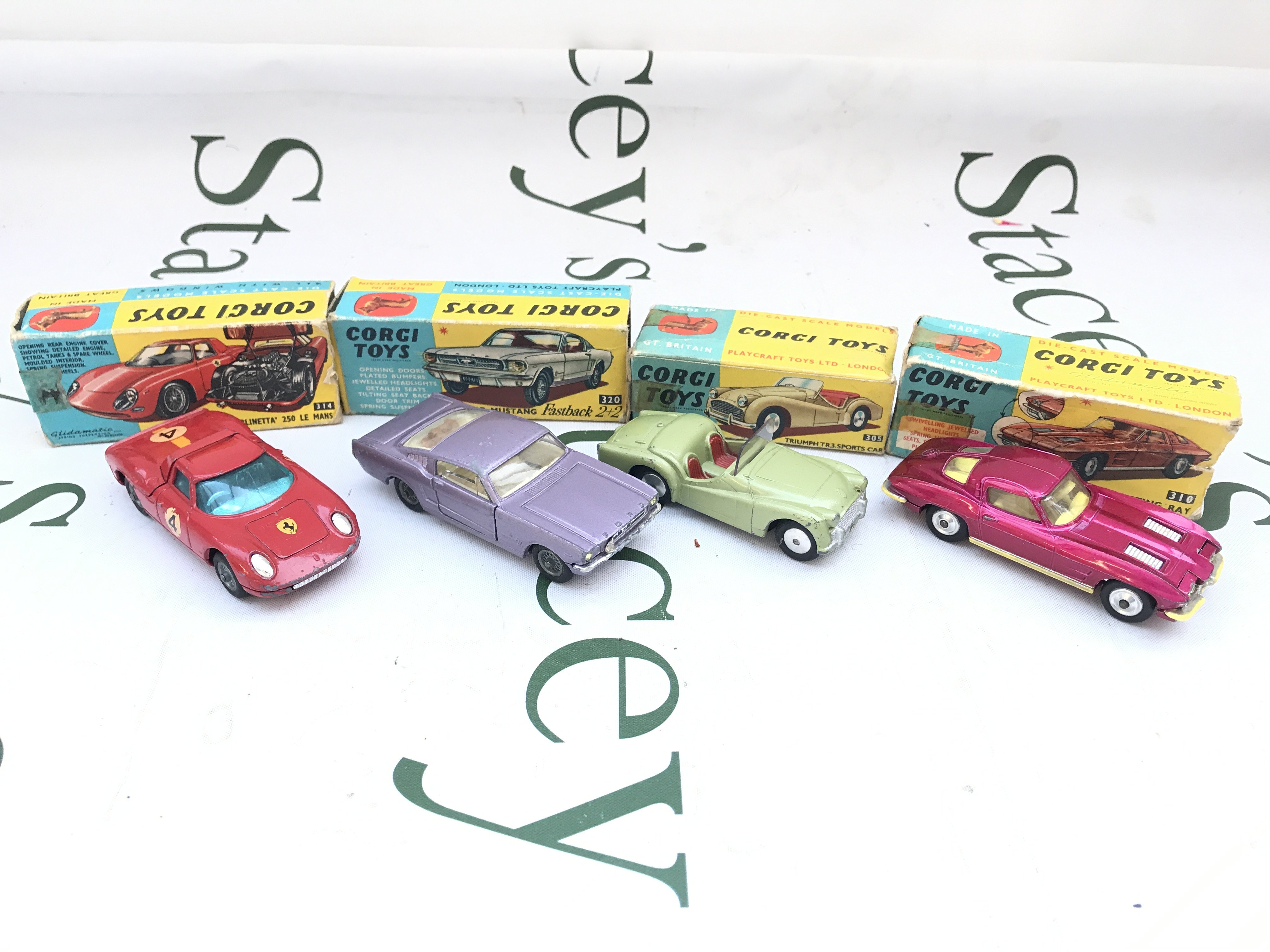 4 Boxed Corgi Vehicles. A Ferrari Berlinetta #314. A Ford Mustang #320. A Triumph TR.3 Sports Car #305 And A Chevrolet Corvette Sting Ray #310. Boxes Are Worn.