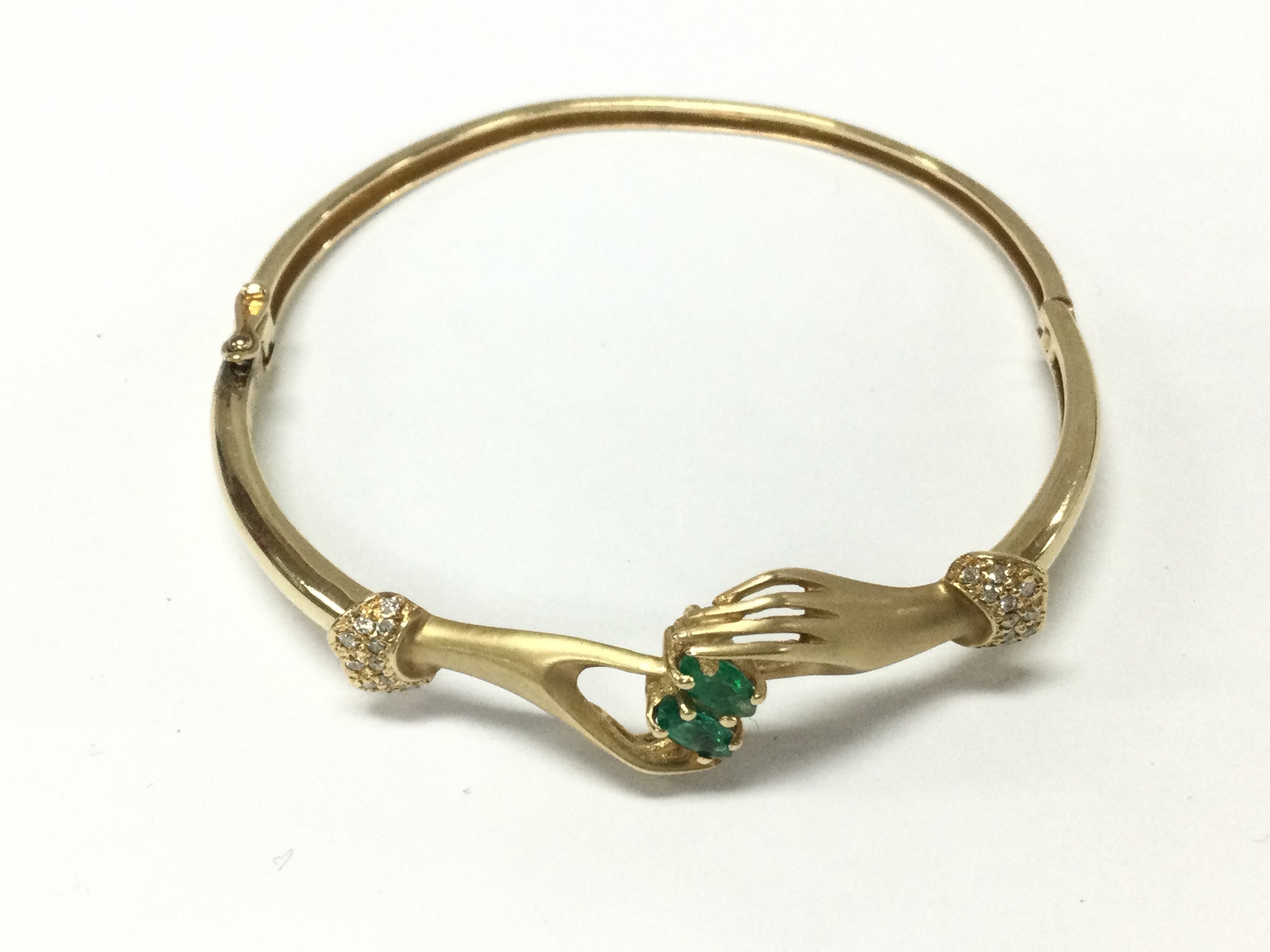 A 14ct gold crossed hands bangle inset with two Colombian green emeralds and pave set diamonds. 9.80g