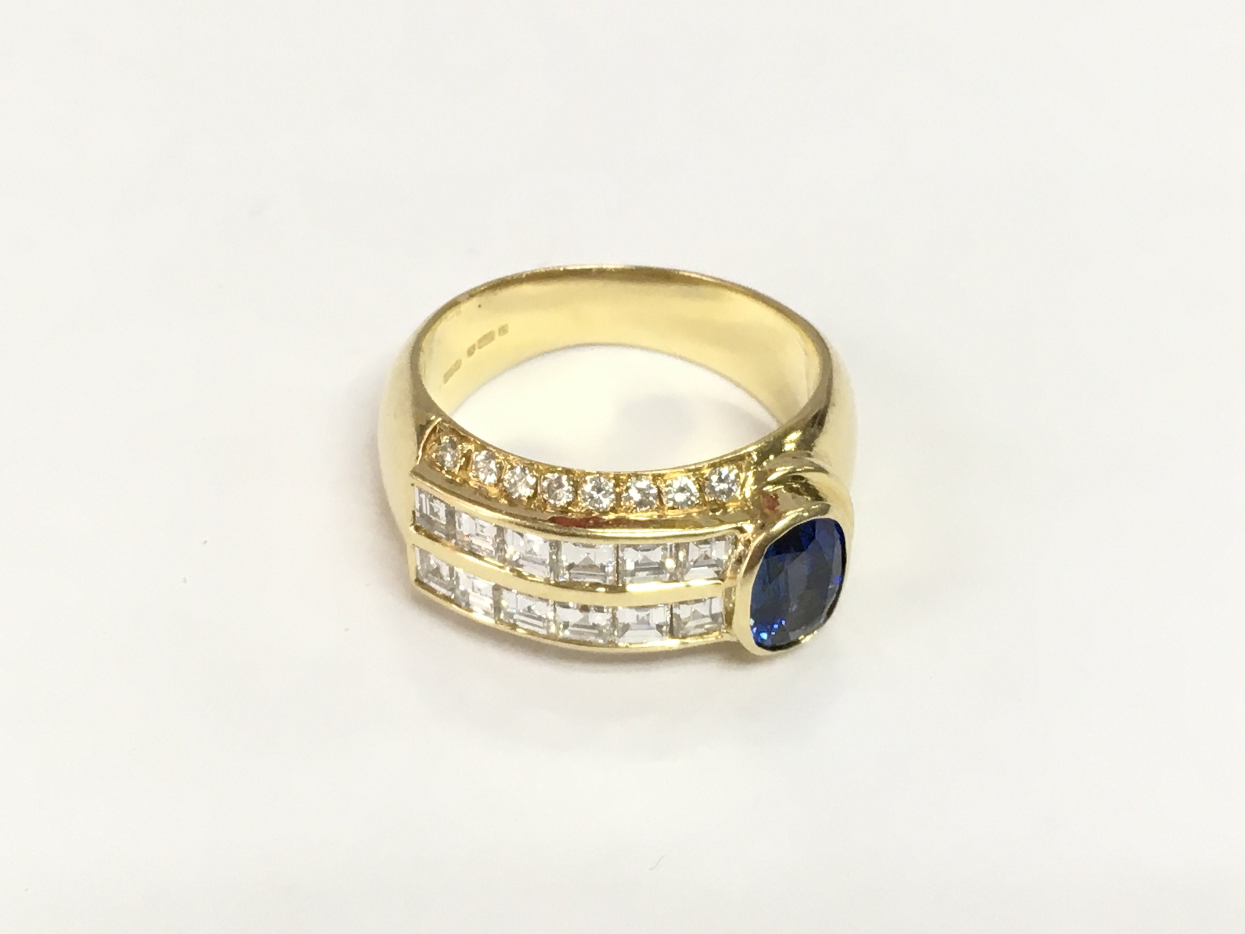 An 18ct yellow gold ring set with an oval sapphire set to one side with square baguette cut and RBC diamonds set to the band. Diamonds approx 1.00ct, sapphire approx 0.70ct. Approx 9.3g and approx size L-M. Shipping category A.