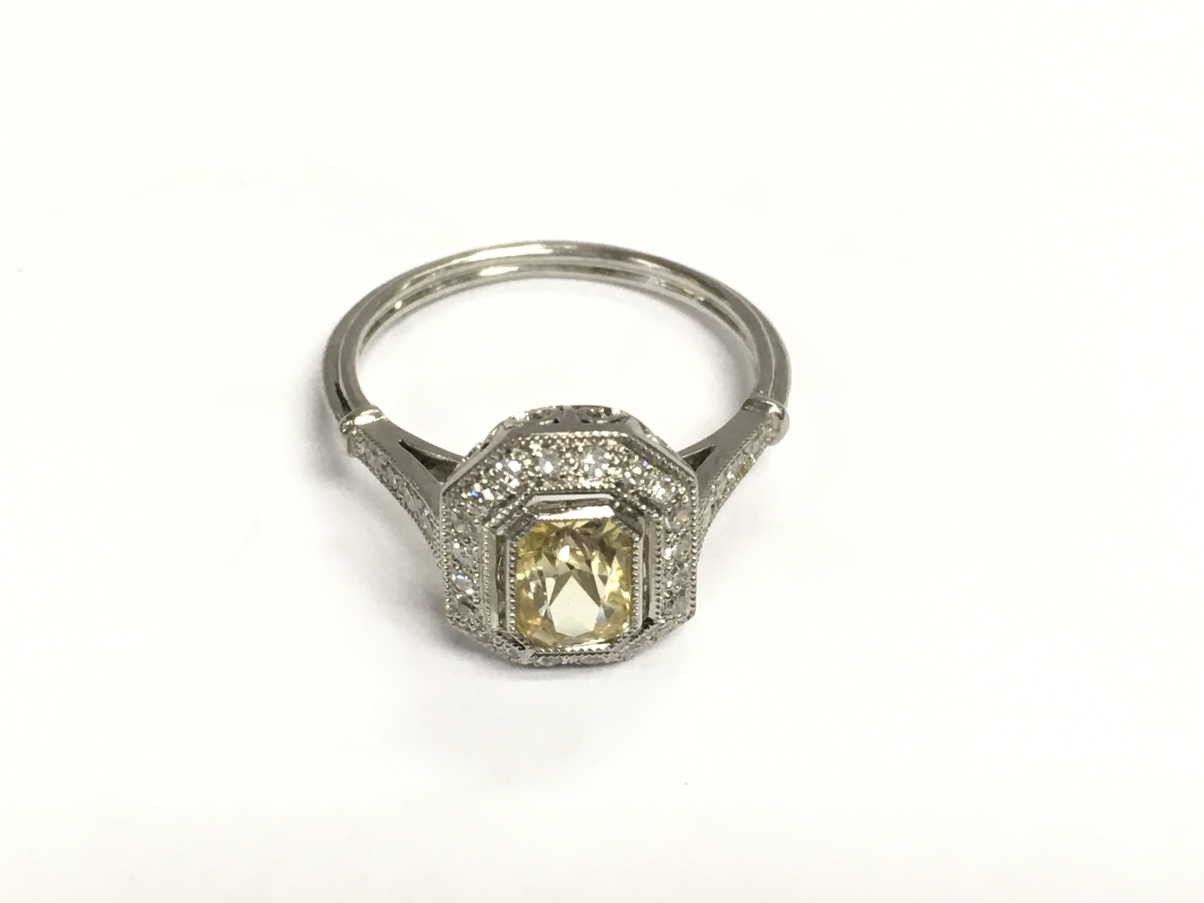 A platinum ring set with a central yellow sapphire surrounded by R/C diamonds with diamond set shoulders and filigree detailing. Yellow sapphire approx 1.24ct and diamonds approx 0.20ct. Approx 3.3g and approx size M. Shipping category A.