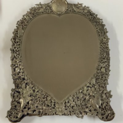 An Antique Victorian hallmarked Sterling Silver dressing table mirror having hand-chased angels, God&#2013266057_s and Goddesses, scroll-work, and floral decoration, with a top engraved cartouche, an original bevelled heart-shaped mirror on a blue velvet backing with easel stand. Made by William Comyns of London in 1887. Height. 51.5cm x 37cm.