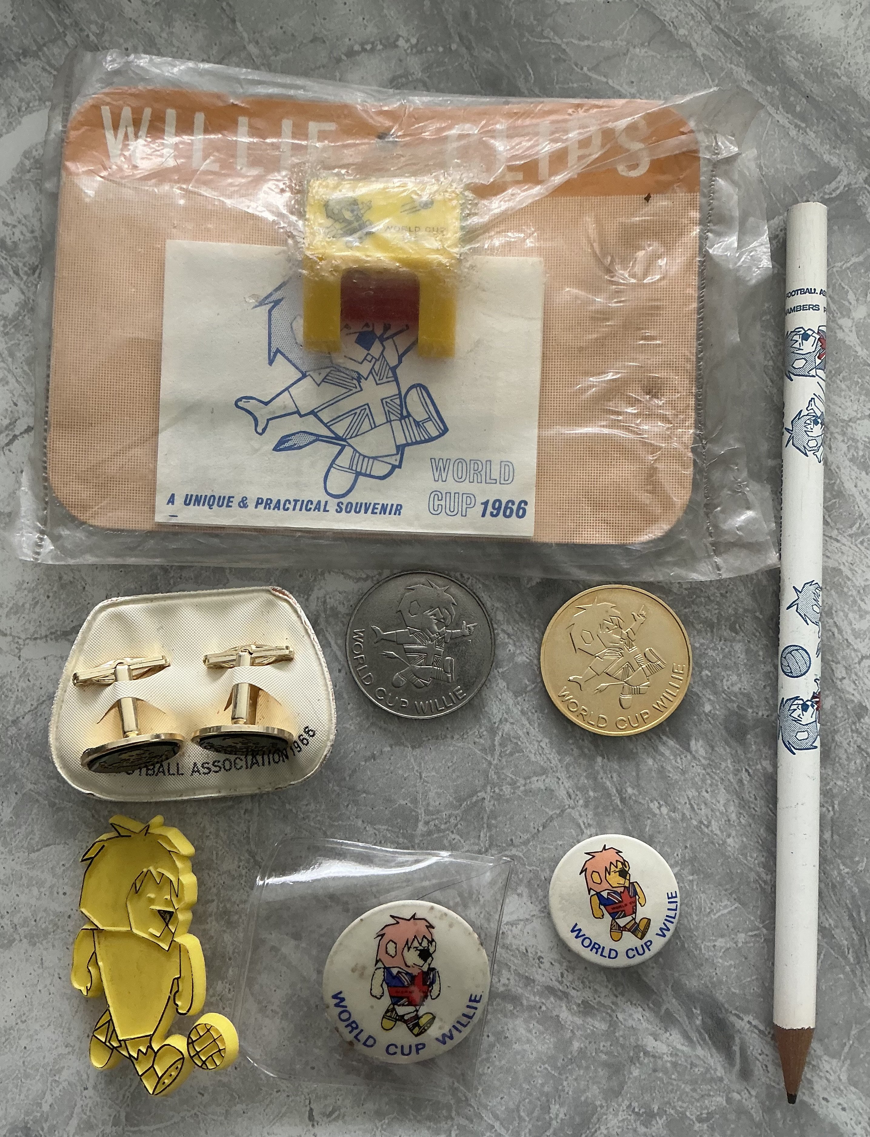 1966 World Cup Willie Football Memorabilia: Pair of cufflinks still attached to Football Association backing, gold and matching silver coin, yellow badge and two different size white ones, pencil and a sealed packet which states Willie Clips and has a plastic clip to attach paper. All 8 items feature World Cup Willie. 
