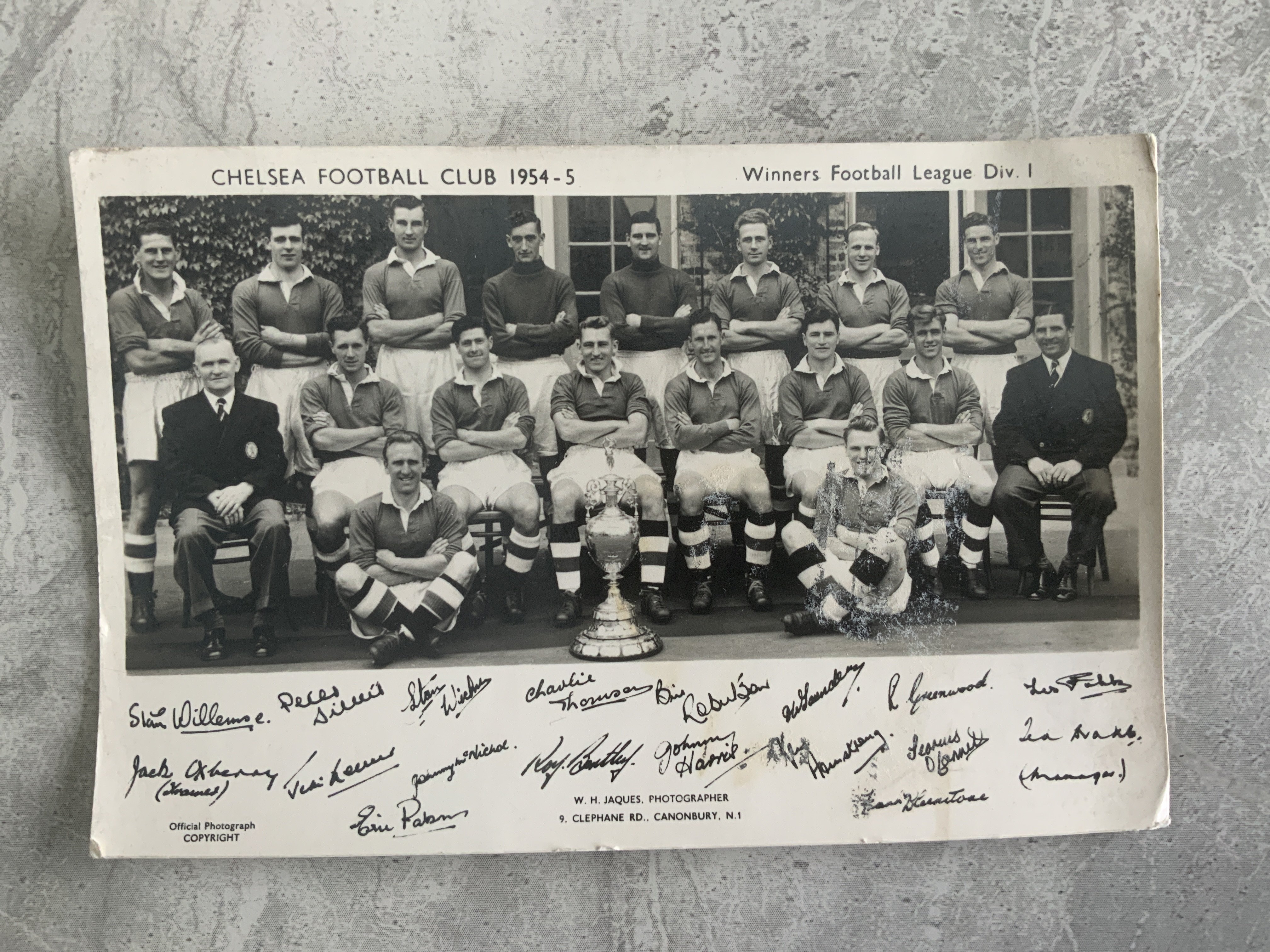 Chelsea 54/55 Framed Photo + Postcard: Division One champions original postcard. Large framed photo of an aerial shot of Stamford Bridge in the period signed to rear board of frame by 8 title winning players. Roy Bentleys autograph is dedicated but Parsons Lewis Smith Willemse Saunders Thomson McNichol etc are not. (2)