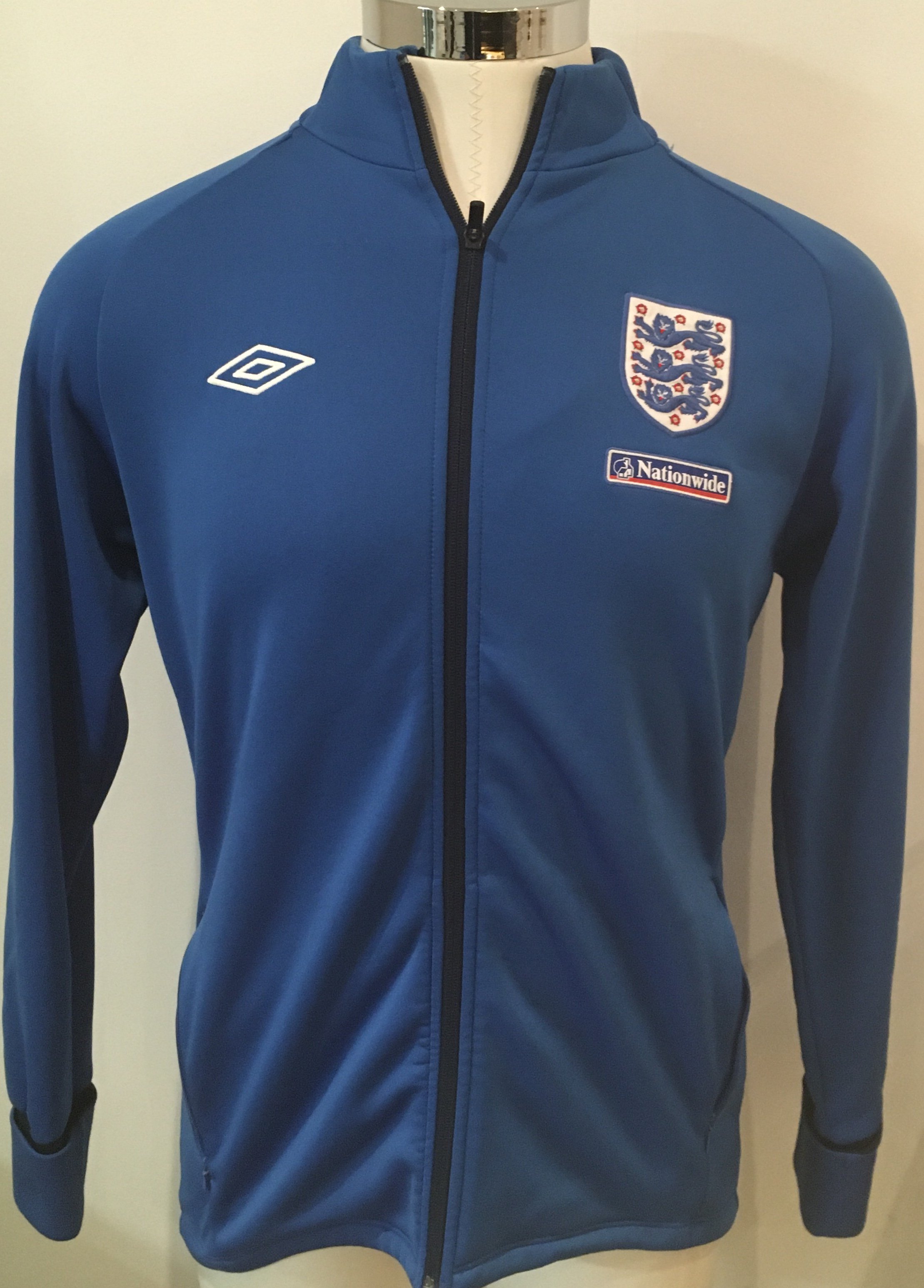 England Signed Football Shirts + Track Suit Top: 3 different era white home England shirts all signed without dedication by Tottenhams Ledley King. A further England shirt is signed by Hoddle and Peter Taylor. These were signed at a Spurs dinner. C/W a lovely blue 90s Nationwide large track suit top with mark to rear and a Score Draw replica England shirt. (6)