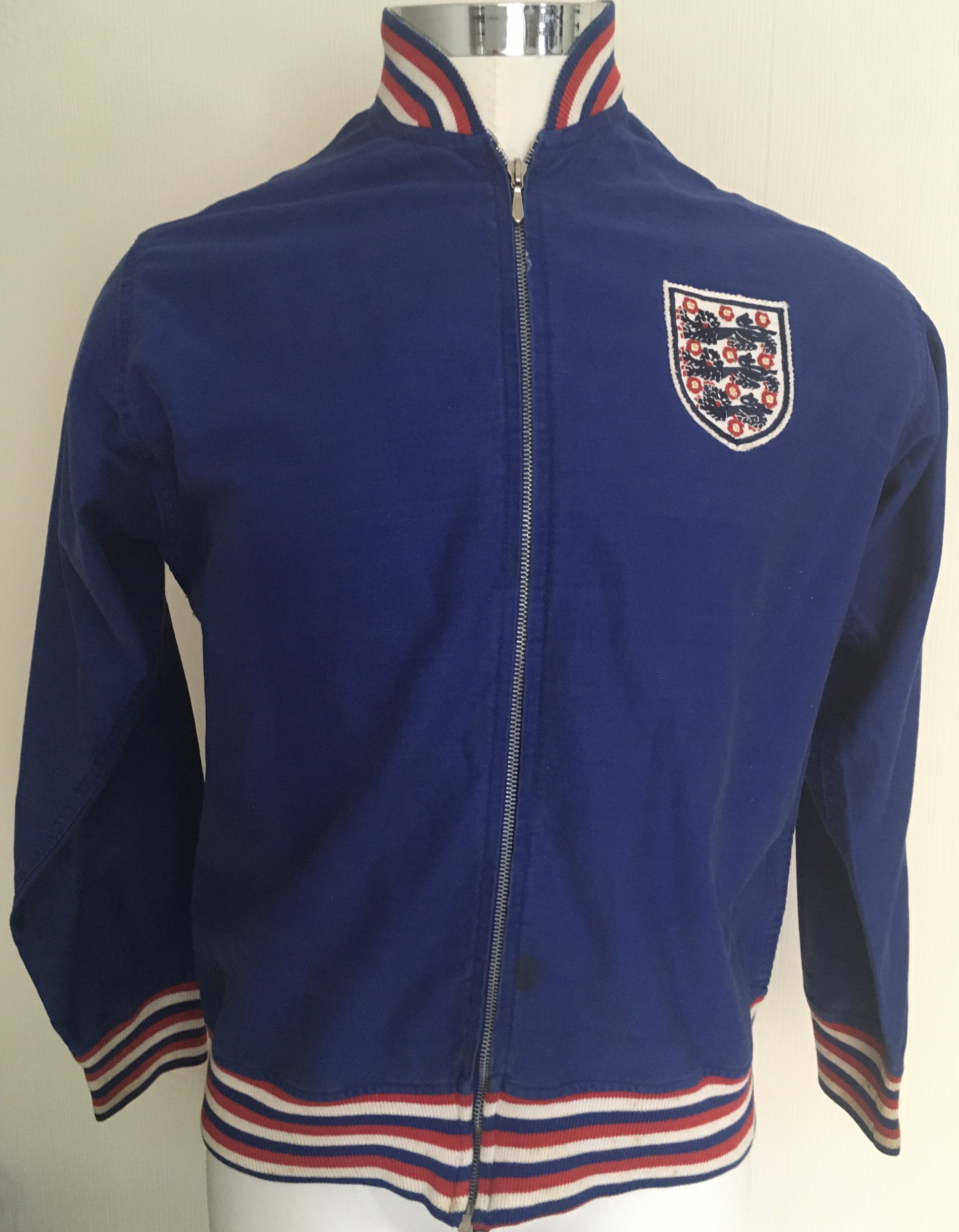 Bobby Moore England Football Tracksuit Top: Umbro top in blue with red and white trim to neck and cuffs. England stitched to rear. Letter of provenance from original vendor is pictured online and states that it was given by Bobby to Mrs Corbett who was a cleaner at Wembley. It came to vendor in a house clearance framed with a magazine picture of Bobby wearing this style top. Now removed from frame with no damage. The make up of this top is correct for around 1970 with embroidery and labelling the same as previous similar items we have had direct from players over the years.