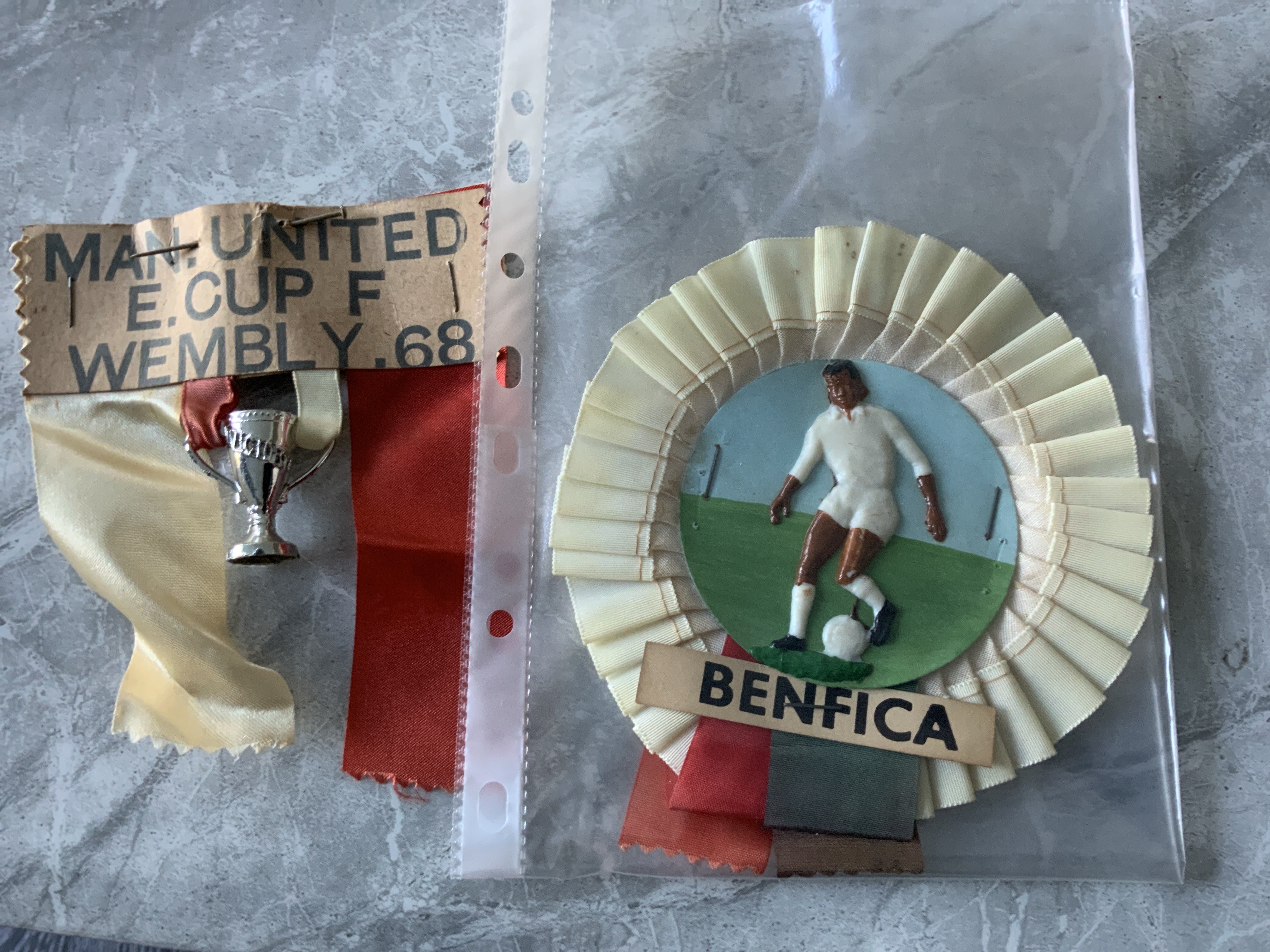 1968 European Cup Final Football Rosettes: Rare Manchester United rosettes with plastic cup hanging from red and white ribbons. C/W Benfica rosette with plastic player in white kit. Nice items. (2)