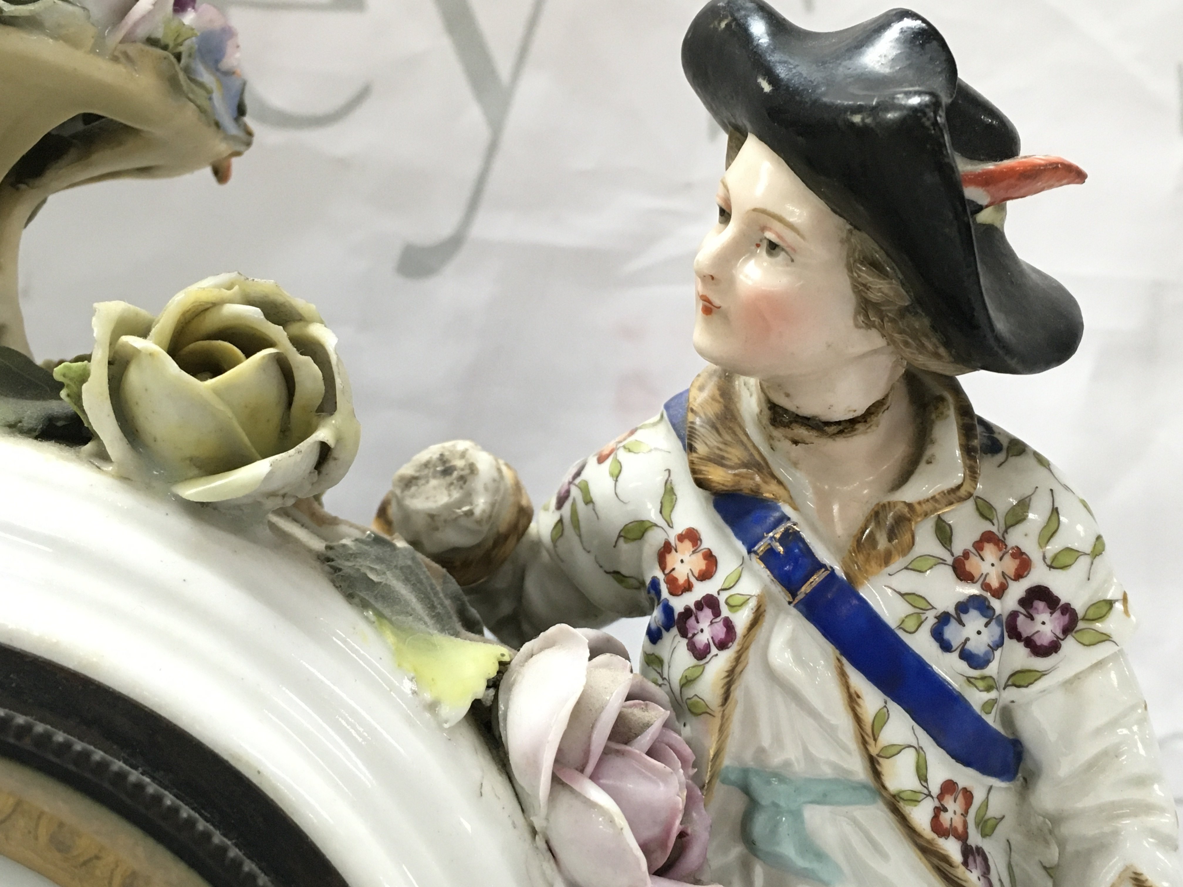 A porcelain clock with raised figures, encrusted with flowers . French movement. Hand missing. Male figures head is damaged. 32cm tall 30cm wide