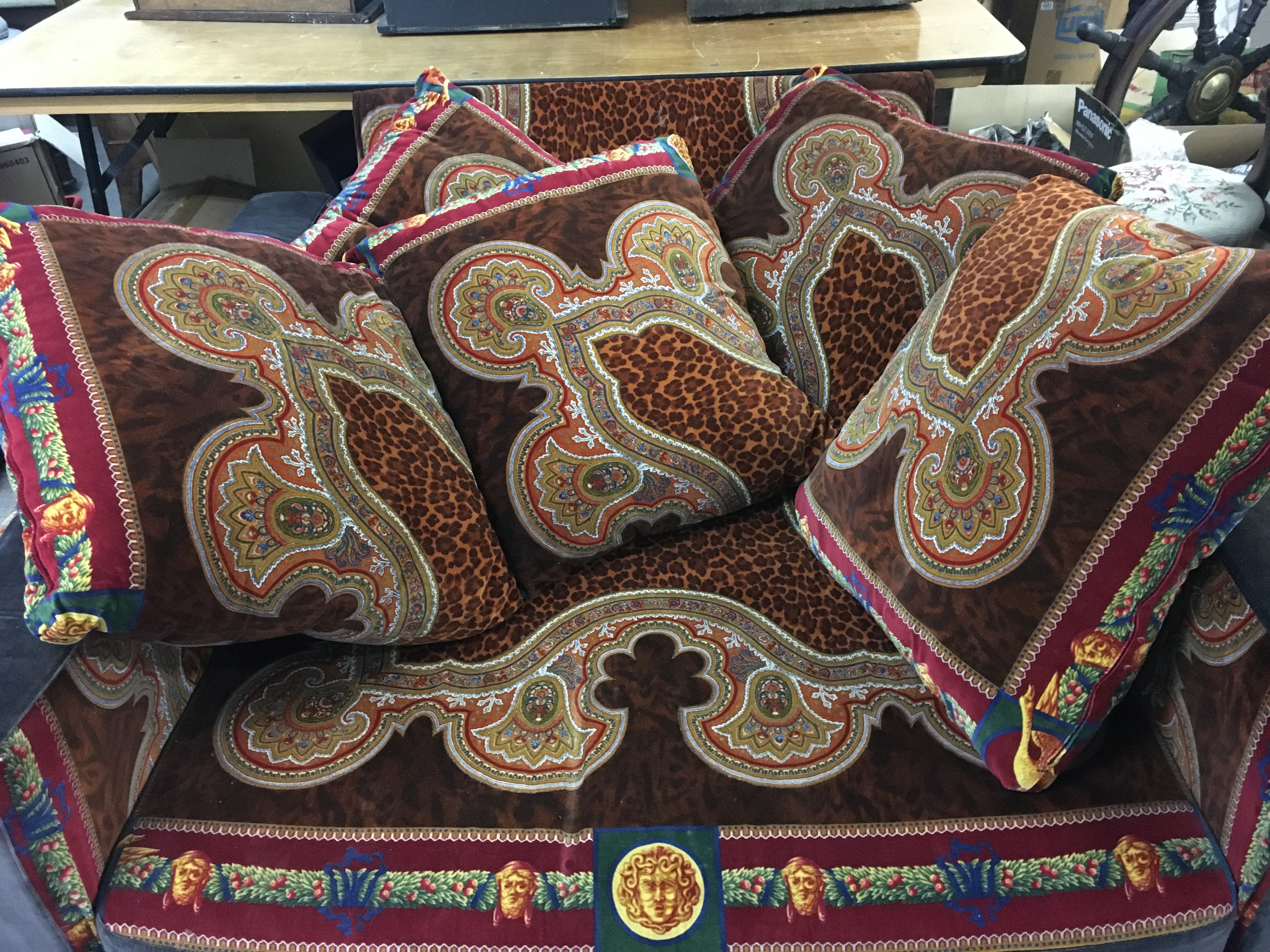 A three piece Versace suite including two sofas, a large chair and ottoman upholstered in a orange, brown , red, green and blue Gianni Versace velvet fabric featuring a leopard and Medusa print. Five cushions and two side fitting coffee tables. Also includes five non matching cushions. Dimensions - Sofas 228cm wide 70cm tall 125cm long , other sofa 225cm wide 60cm tall 118cm deep. Chair 130cm wide 75cm tall, 108cm long. Ottoman 102cm wide 36cm tall 100cm long. Side tables 65cm wide 26cm tall.