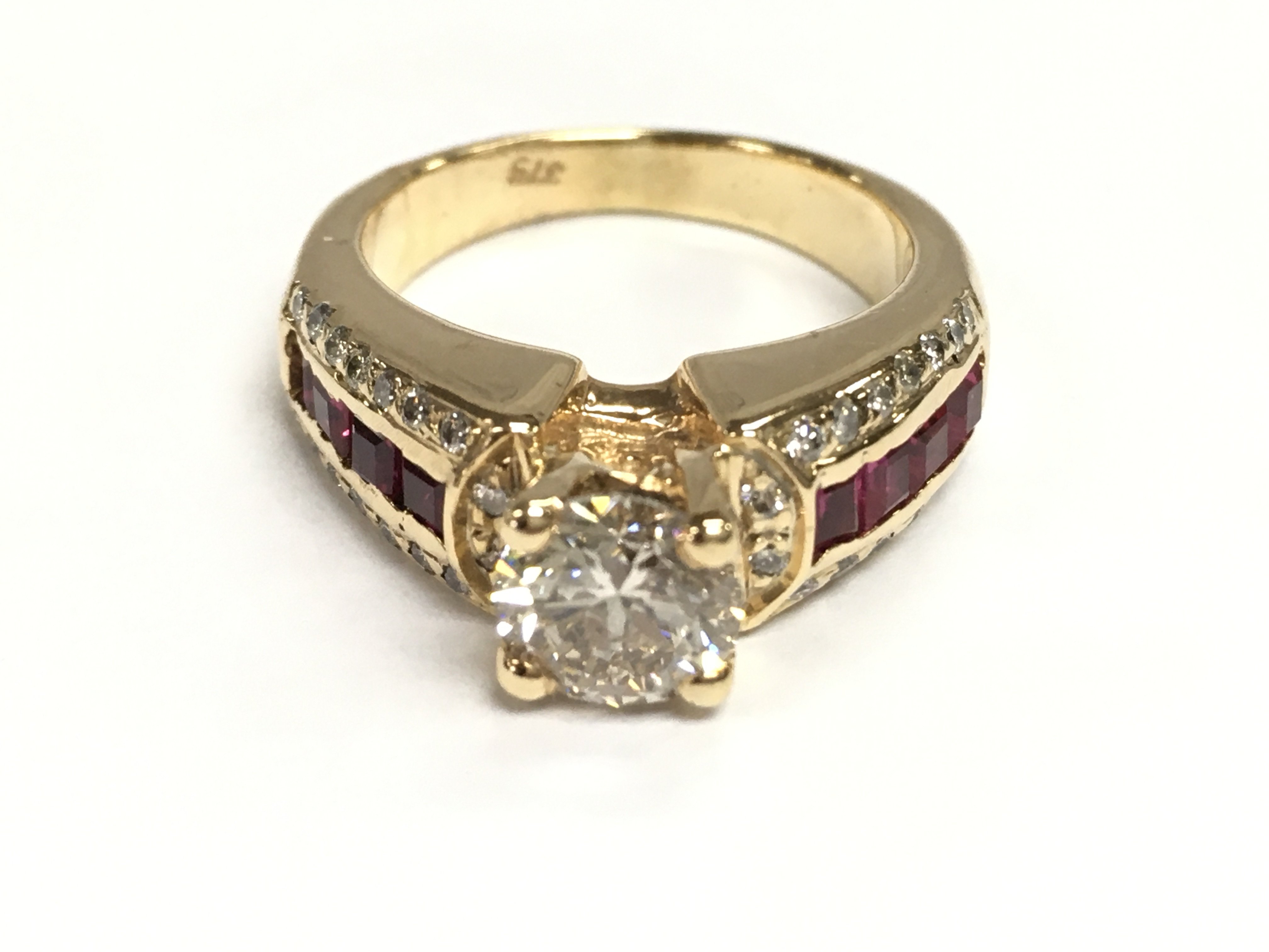 9ct gold ring set with a raised central diamond with further diamonds and red stones on the shoulders. Central diamonds 1.01ct and other diamonds Approx 0.09ct Size M
