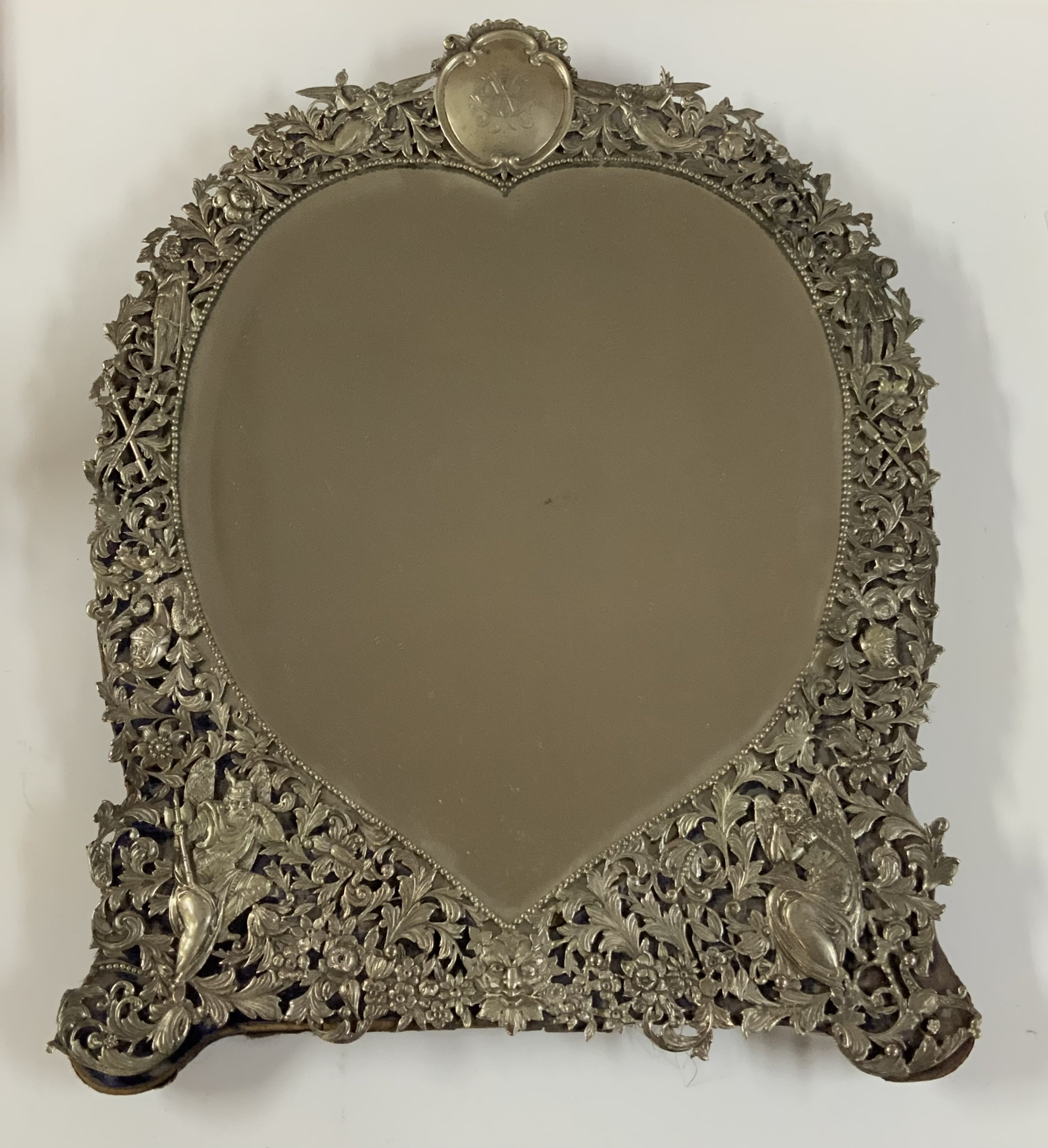 An Antique Victorian hallmarked Sterling Silver dressing table mirror having hand-chased angels, God&#2013266057۪s and Goddesses, scroll-work, and floral decoration, with a top engraved cartouche, an original bevelled heart-shaped mirror on a blue velvet backing with easel stand. Made by William Comyns of London in 1887. Height. 51.5cm x 37cm.