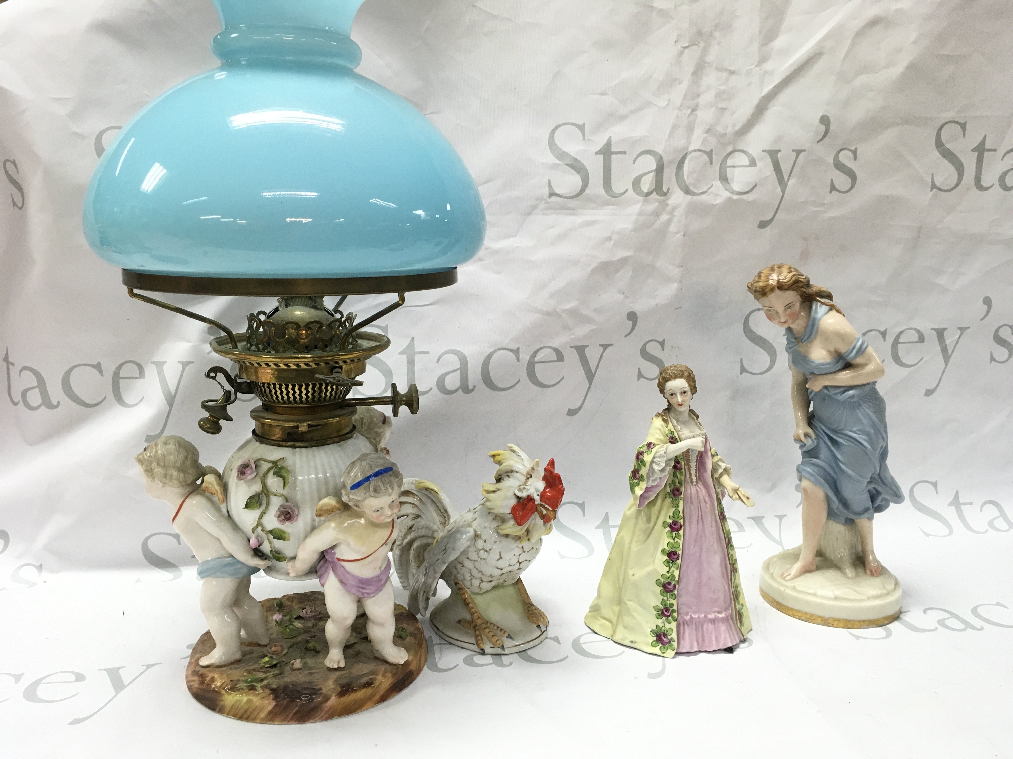 Porcelain figures including Worcester against the wind , Madame du Barry (with restoration) Mesissen cherub figure group oil lamp with a light blue glass shade, German rooster figure. This lot cannot be posted