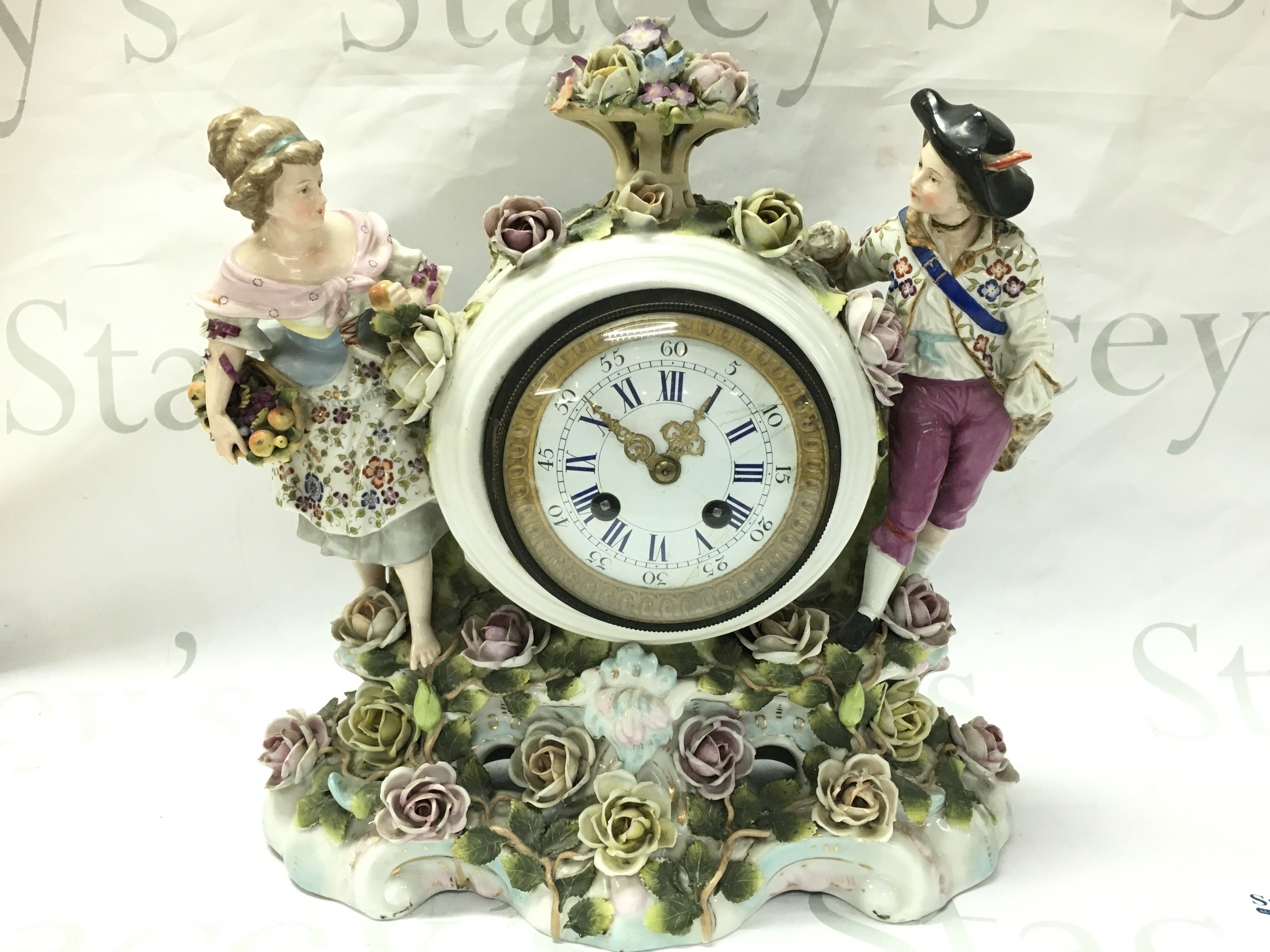 A porcelain clock with raised figures, encrusted with flowers . French movement. Hand missing. Male figures head is damaged. 32cm tall 30cm wide