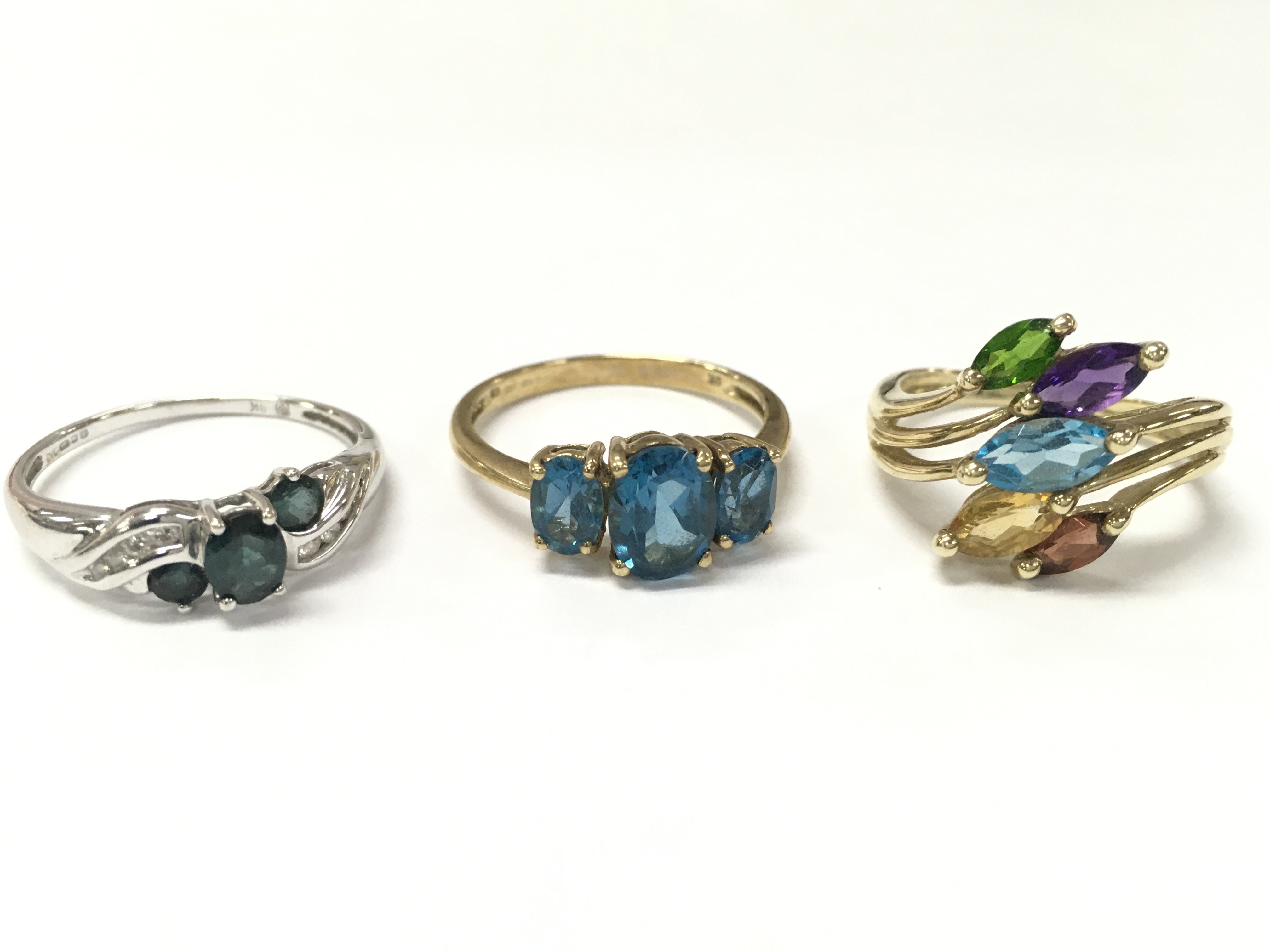 Three 9ct gold rings including a teal sapphire and white stone set white gold ring size R, Blue topaz trilogy ring size R, multi gem ring size O. 8.2g total weight. Postage category A