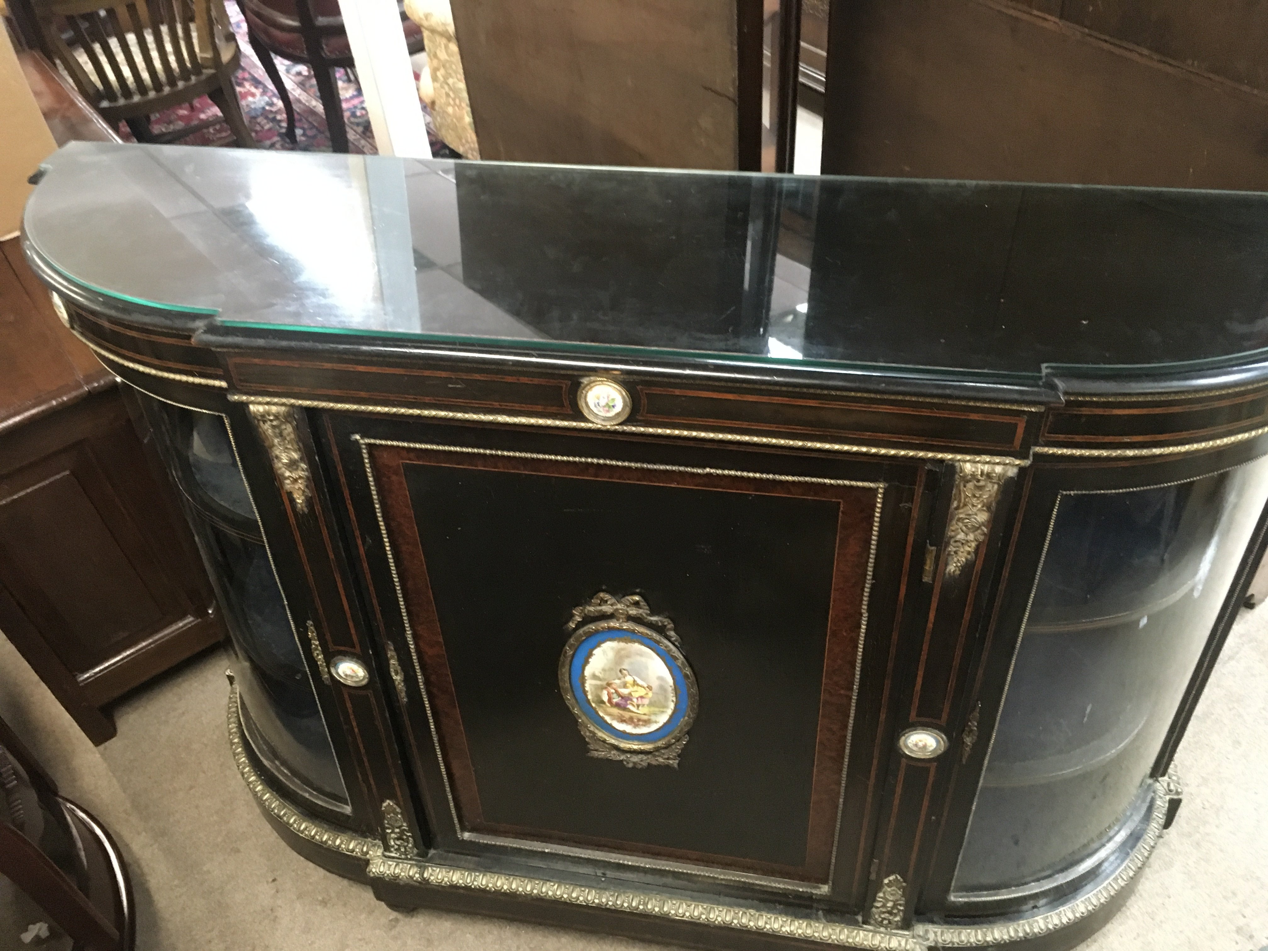 A Victorian ebonised credenza with two glass doors on either side with shelves for storage, gold ormolu trim and plaques ,porcelain hand painted panels of floral imagery and in the middle a classical figures fishing scene ,with a glass top. A foot is loose and needs reattaching. 150cm wide 98cm tall.