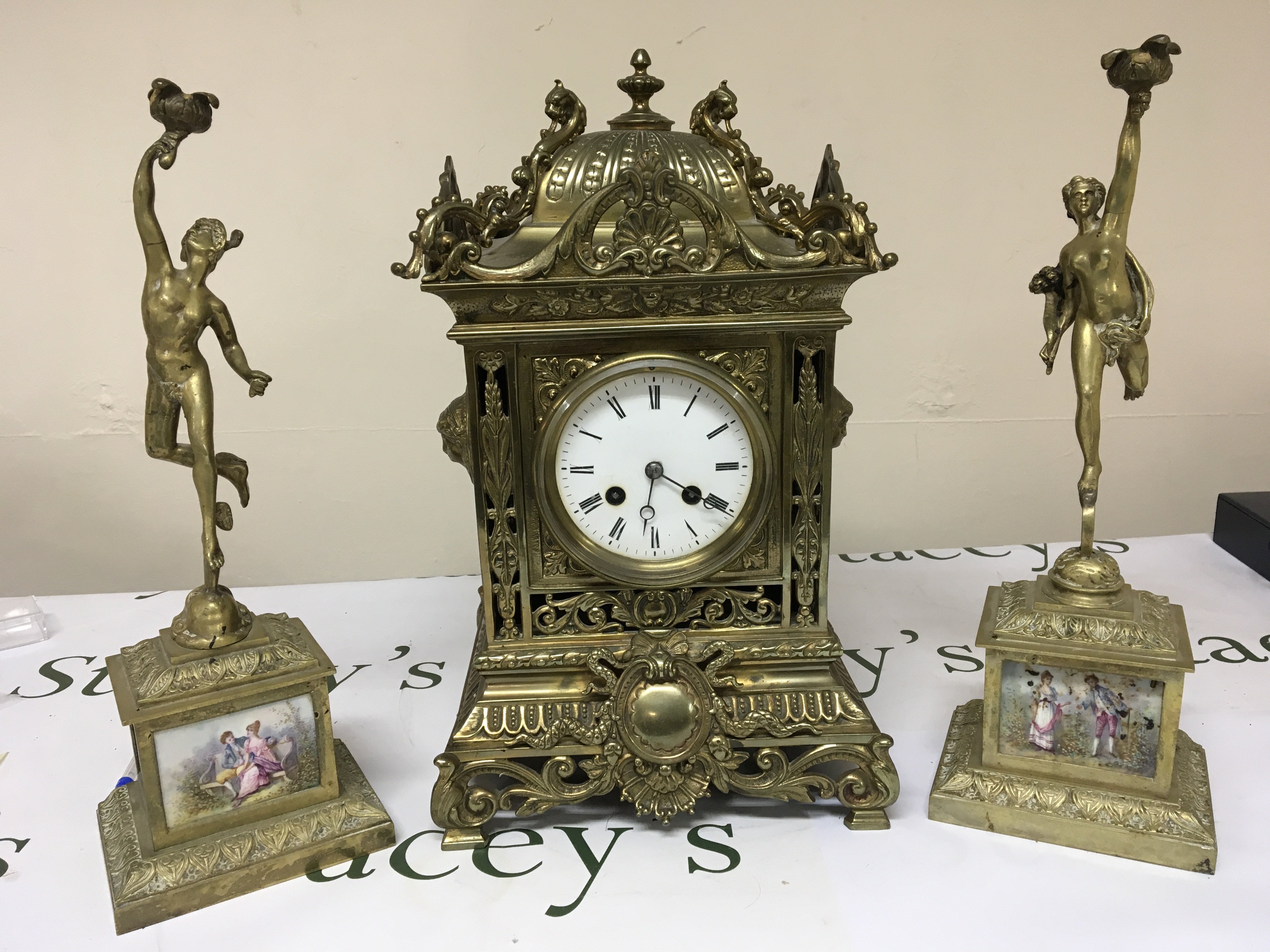 A late 19th Century brass cased clock garniture the elaborate clock case with raised scrolls. The side ornaments with figures of Mercury and set with hand painted porcelain panels. decorated with figures. The movement striking on a bell and with pendulum.