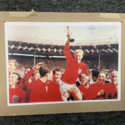 Bobby Moore Signed England Football Print: An A4 print of the famous picture of Moore being held aloft holding the World Cup. Hand signed in blue biro Best Wishes with no dedication. Mounted on board.