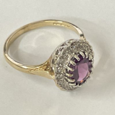 9ct yellow and white gold dress ring set with a purplish red rhodolite garnet and an R/C diamond halo. Oval cut garnet 0.60ct. Diamonds 0.12ct. Postage category A.
