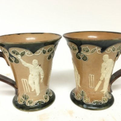 Two Matching Lambeth Doulton stoneware mugs decorated with raised cricket figures impressed marks to the base one with a small chip. Height 12.5cm