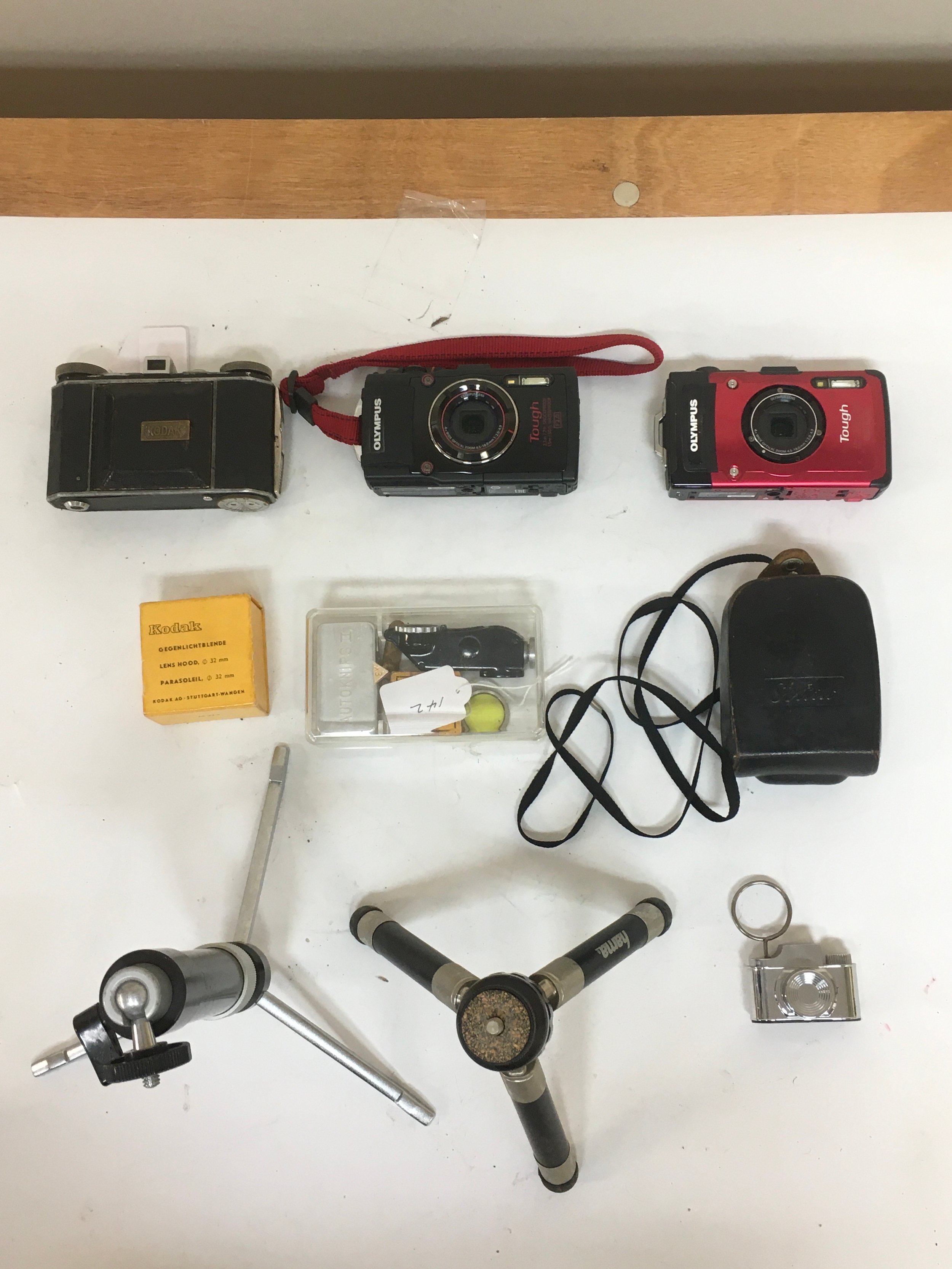 Assortment of cameras, Kodak Compur, Olympus Tough digital Cameras. Black and red colour variations with strap. Kodak lens hood with box. Box with array of lens equipment and view finder, additional view finder. CDS Sixtar with pouch carrier. 2 camera stands. Camera key ring.