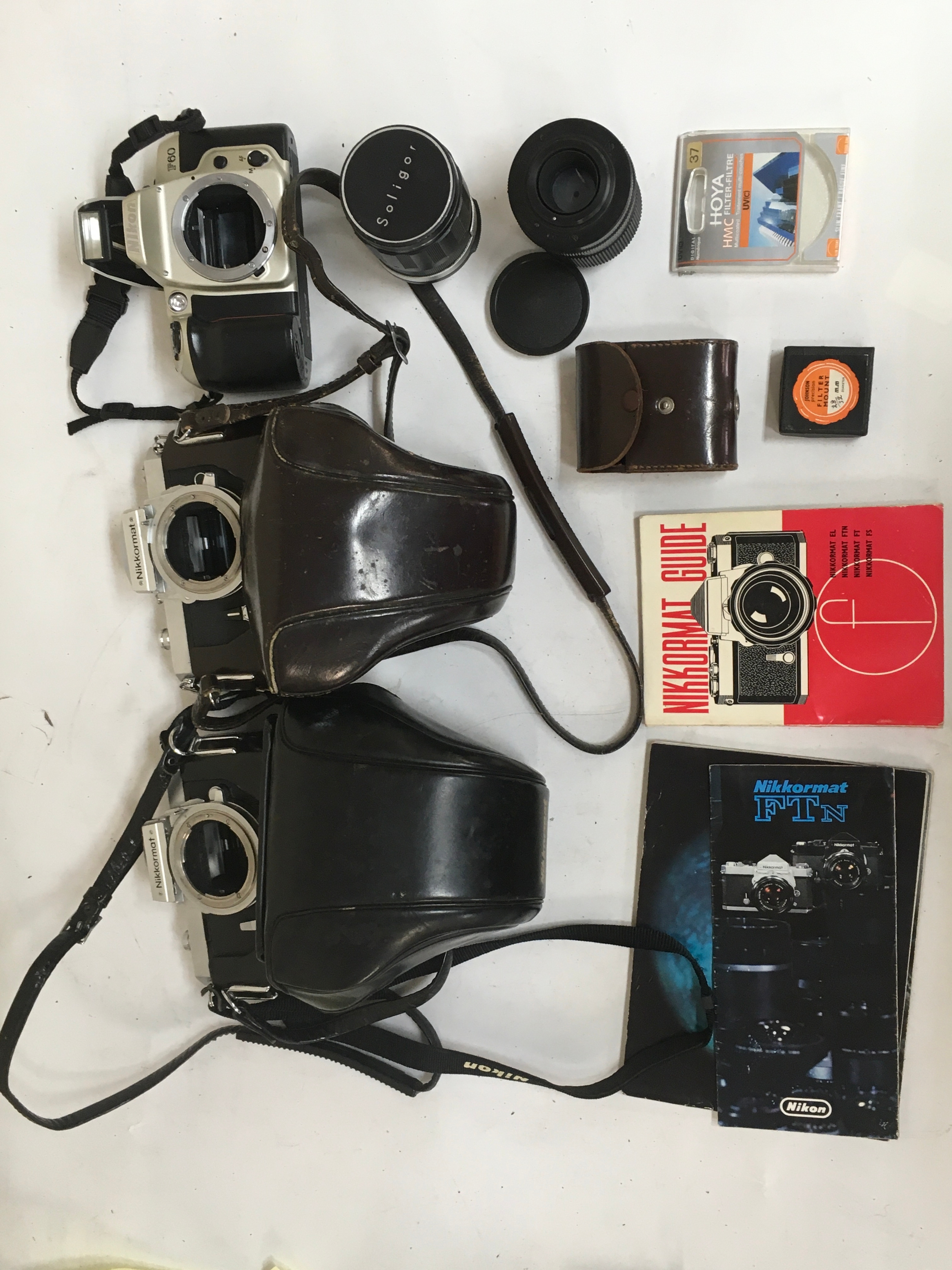 10 items.  3 Cameras. 2 Nikkormat Nikon Cameras with holster / carriers. No/  missing lenses. 1 Nikon F 60 camera with flash, no/ missing camera lens. Miscellaneous camera accessories 2  Soligor Camera lenses with cover. An assortment of Nikkormat Guides for FTN and other modals. Filter Mount. HOYA Filter.  1 small leather pouch for various Retina camera parts.
