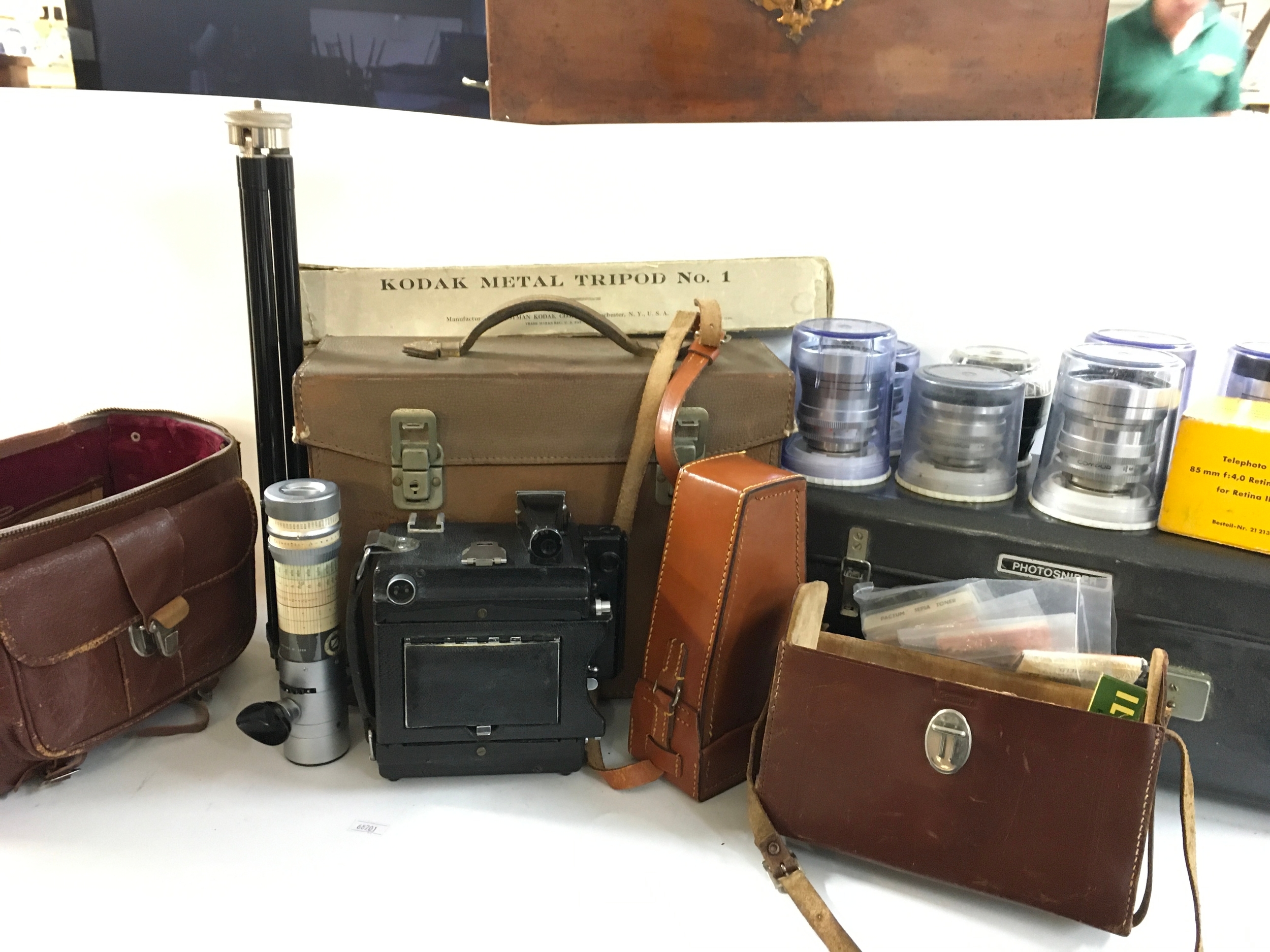 A collection of vintage photography gear including a Kodak Metal Tripod, a Press camera, a vertical optical rangefinder attachment, a carrying case, film boxes and a range of different lenses. 