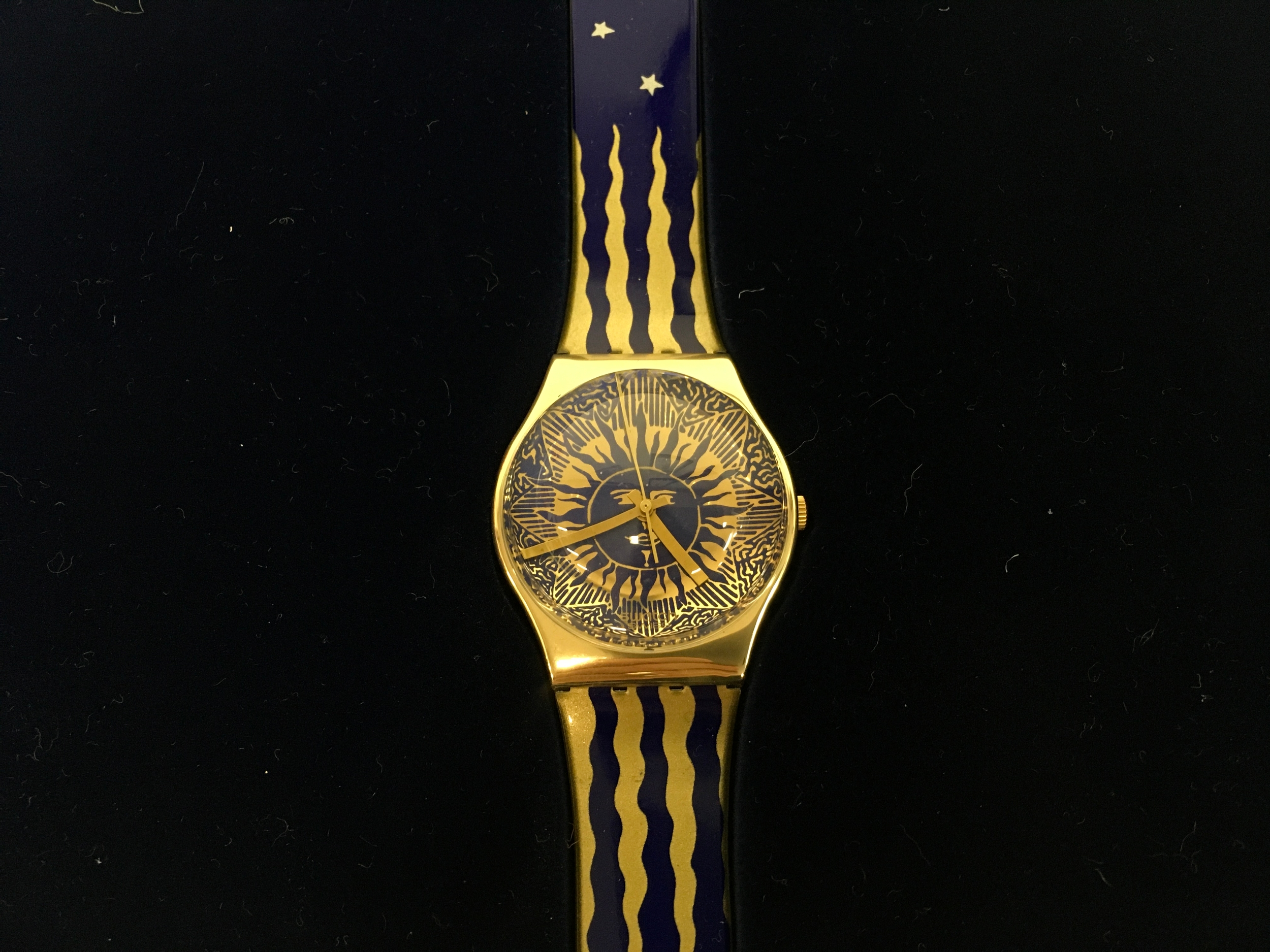 1991 Swatch Gent "Hocus Pocus" X-Mas Special (GZ122) watch, packaged in its original blue metallic case featuring a celestial star print. The watch features a blue plastic strap with gold sunray and silver star patterns, paired with a gold and blue dial. Limited to 14999 pieces worldwide, it was designed in collaboration with Byron Glaser and Sandra Higashi.