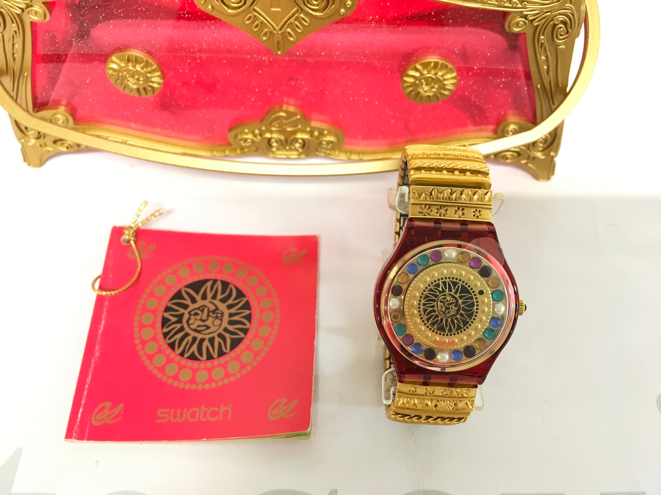 Swatch "Xmas by Xtian Lax" (GZ140) wristwatch from 1994, designed by Christian Lacroix, comes in its original special presentation box. The watch features a gold-toned flex bracelet and a unique face design. It is housed in a decorative, transparent red and gold jewelry-style presentation box.