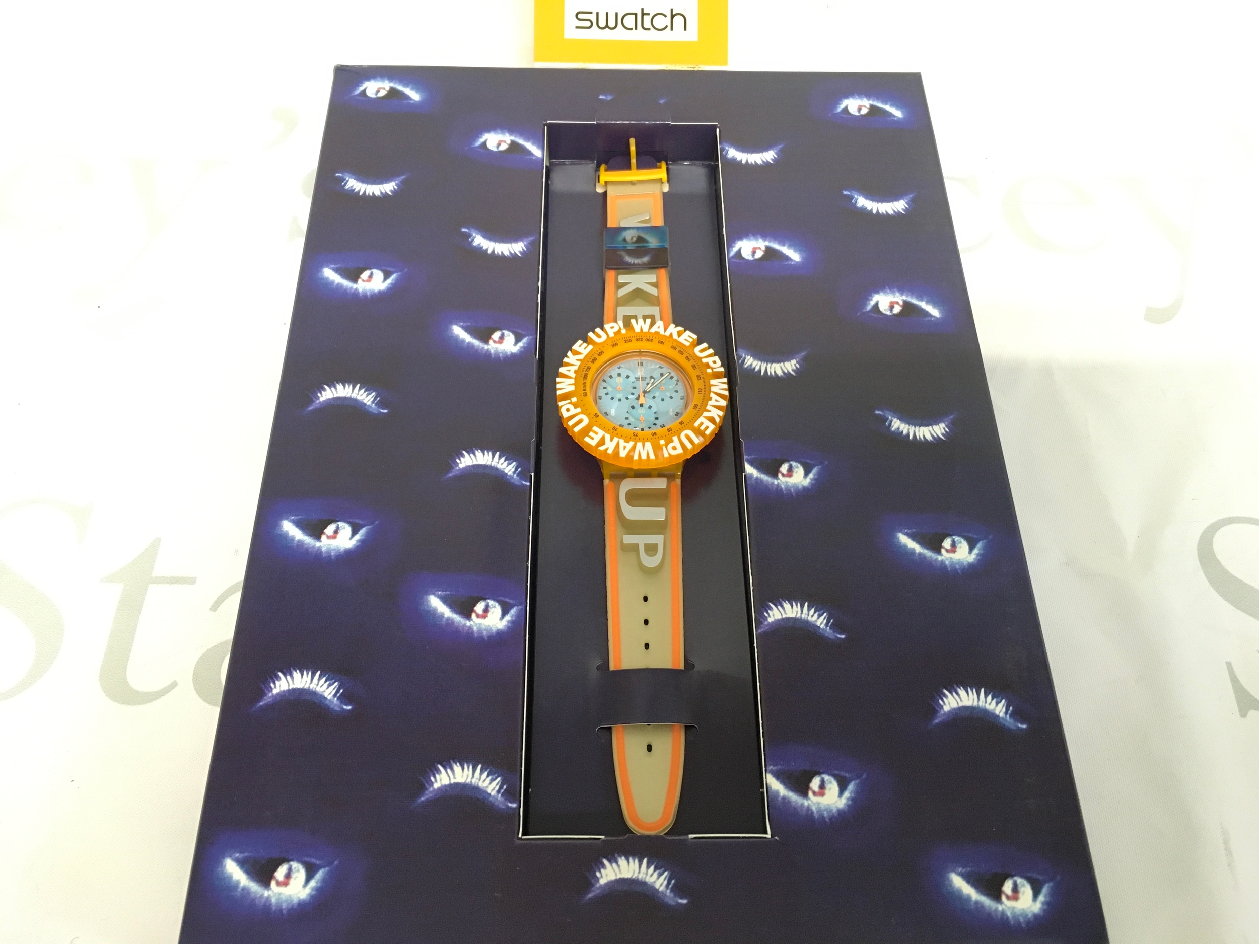 Swatch Aqua Chrono "Wake Up" (SBZ104) watch, a 1998 special edition designed by Spike Lee. It features a distinctive 45mm diameter orange bezel with "WAKE UP!" text and a light-colored strap with orange accents.