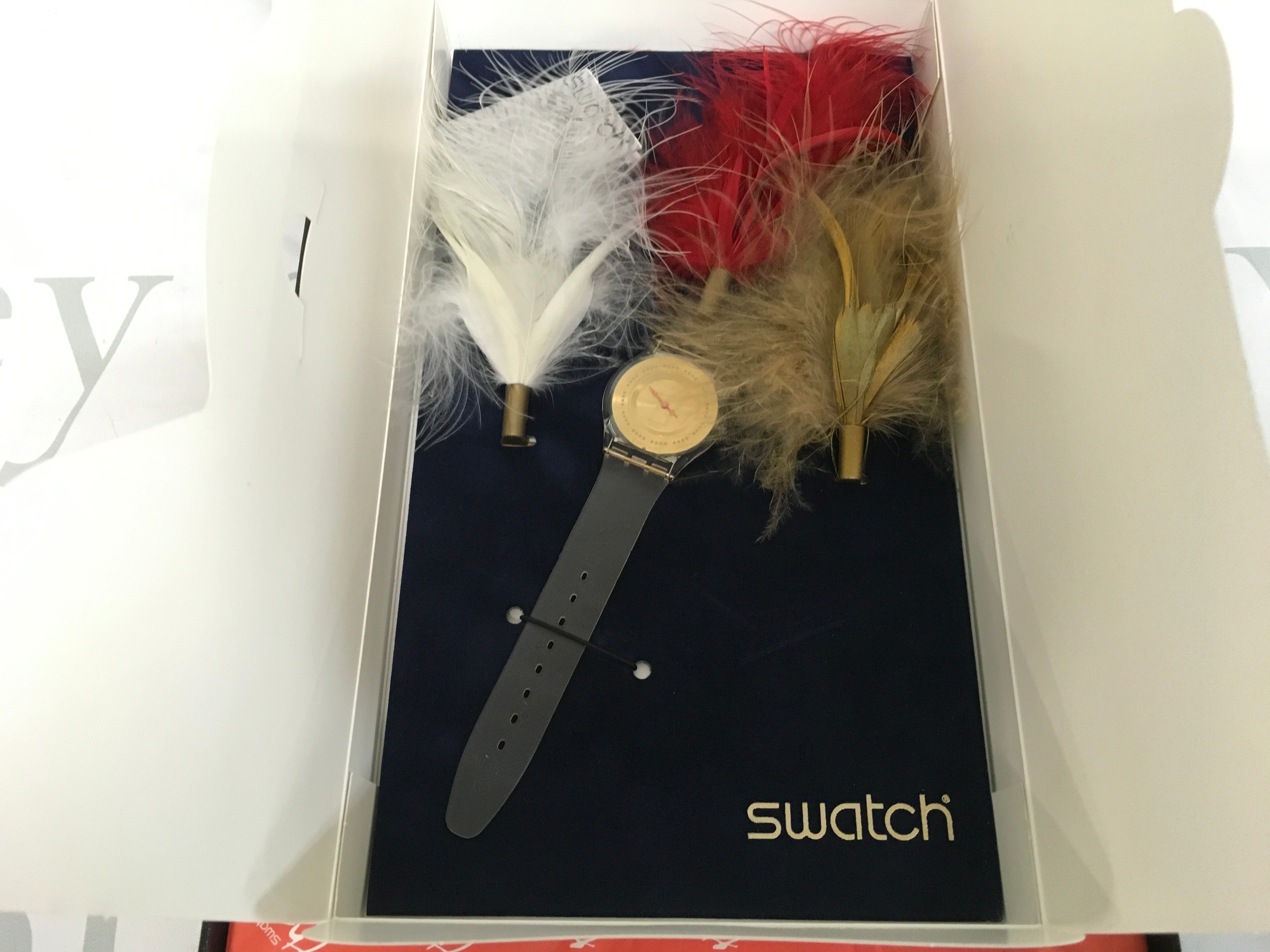 Swatch "Plume de Fete" (SFK136Pack), a limited edition watch released for the 2001 Christmas Special. It features an ultra-thin Skin Classic watch with a gold-colored dial, paired with decorative white, red, and gold feathers. The set was limited to 9,999 pieces worldwide