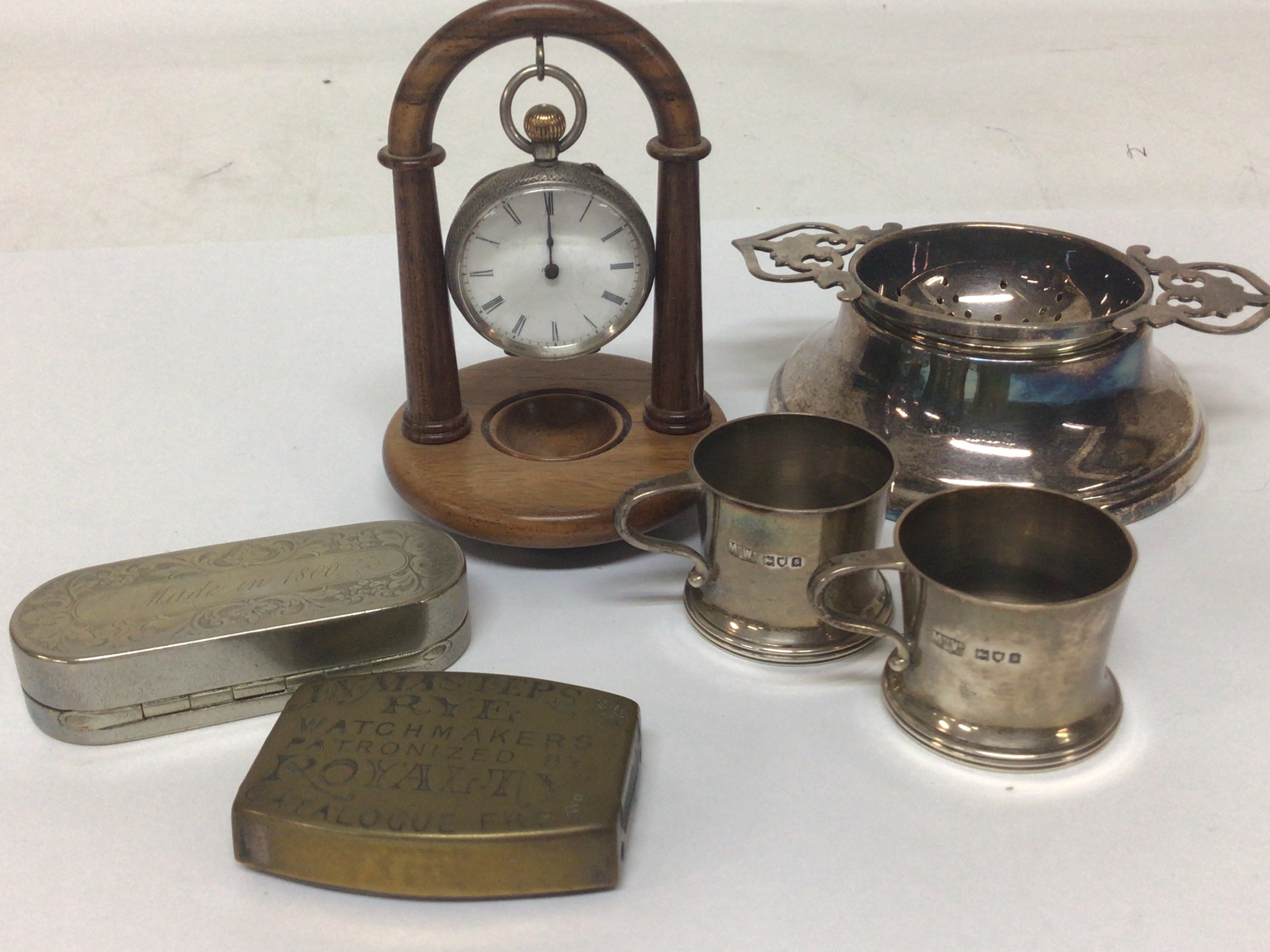 A Victorian watchmaker's advertising vesta case a metal snuff box with inscription made in 1800 a small silver button wind pocket watch on a turned wood stand two miniature silver mugs and a tea strainer with silver base. 
