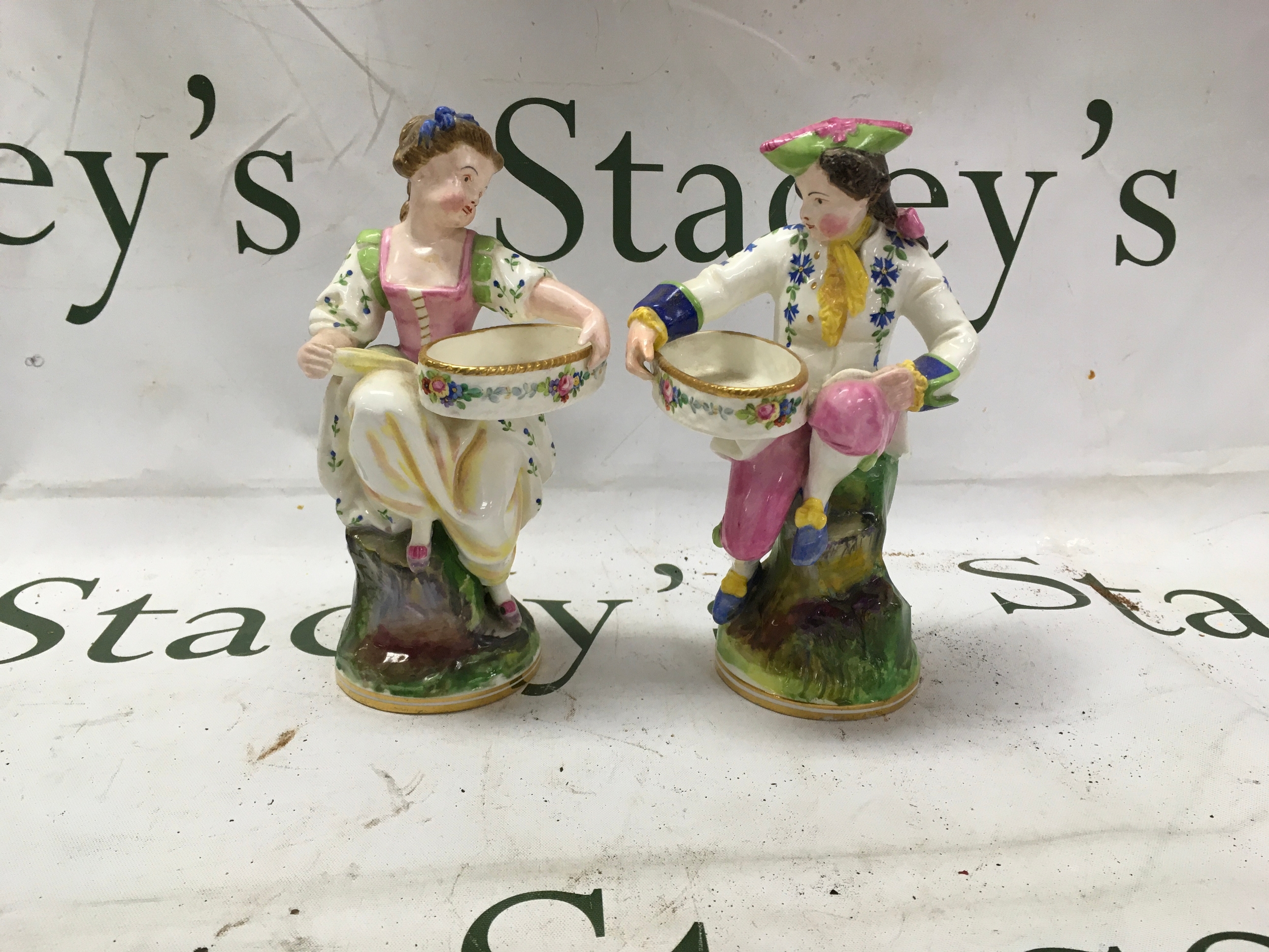 Pair of Minton basket carrier sweetdishes, designed as a amn and woman in 18th c dress, the man has professional restoration to his thumb, marked to the base withpuce Minton marks. 18 cm high.