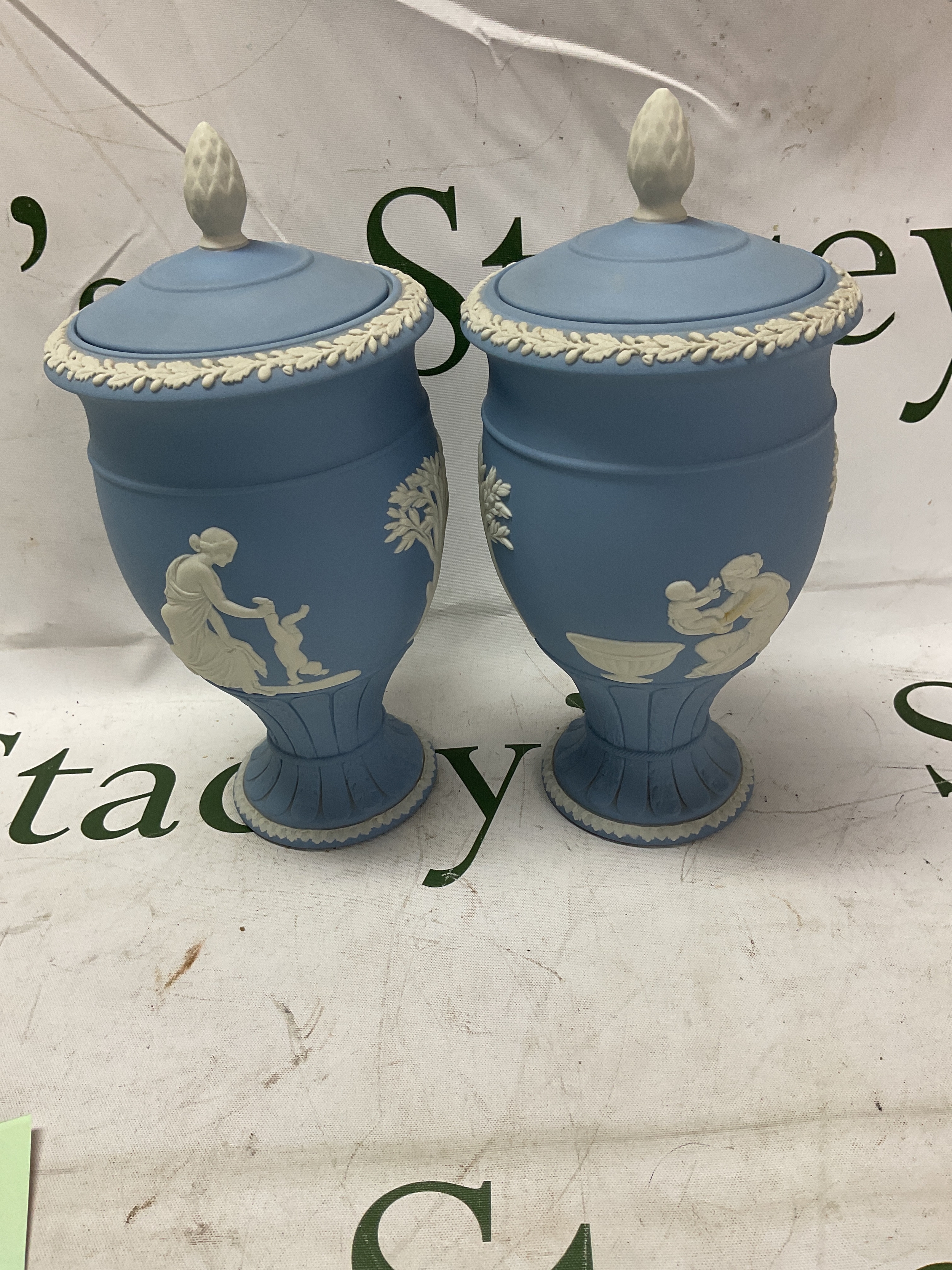 Wedgwood pair of lidded urns in traditional blue, depicting Achilles mother and Moses mother with child, pineapple finial to the lids. 22cm H