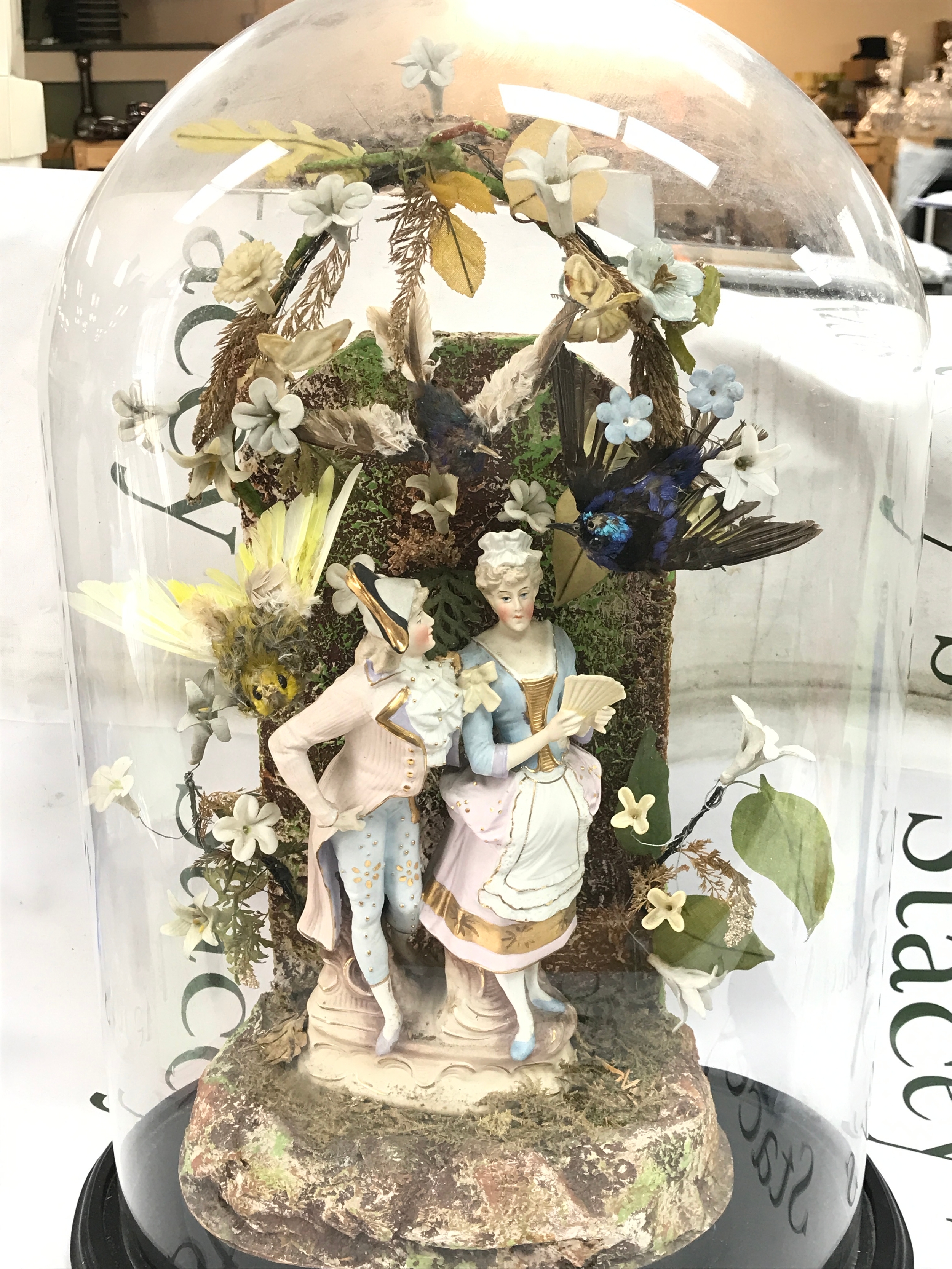 A 19th-century continental bisque figural group of an 18th-century style lady and gentleman surrounded by flowers and foliage beneath a glass dome on an ebonised wooden base. 52cm tall, 38cm wide, 22cm long 