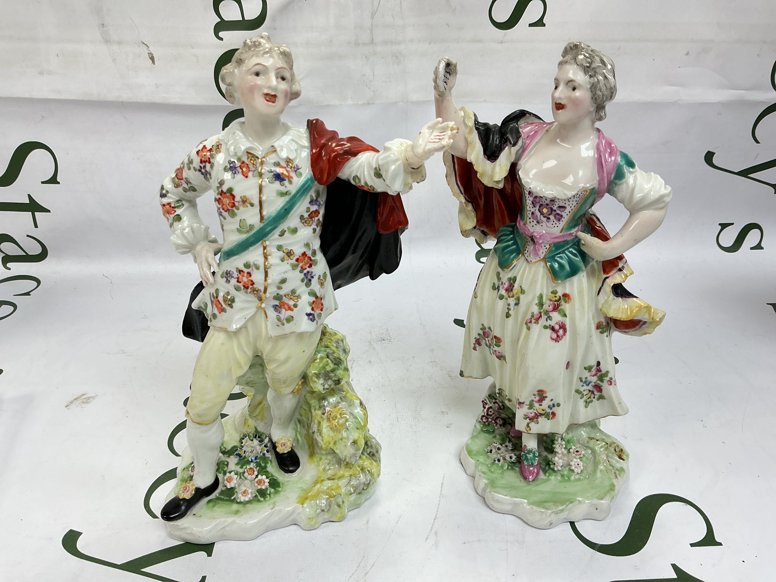 A pair of early 19th century Derby figures of singers, both with one hand on a hip and the other raised holding a song sheet, the couple stand on flower strewn rocky bases, some chips and restoration. Approx height 21cm.