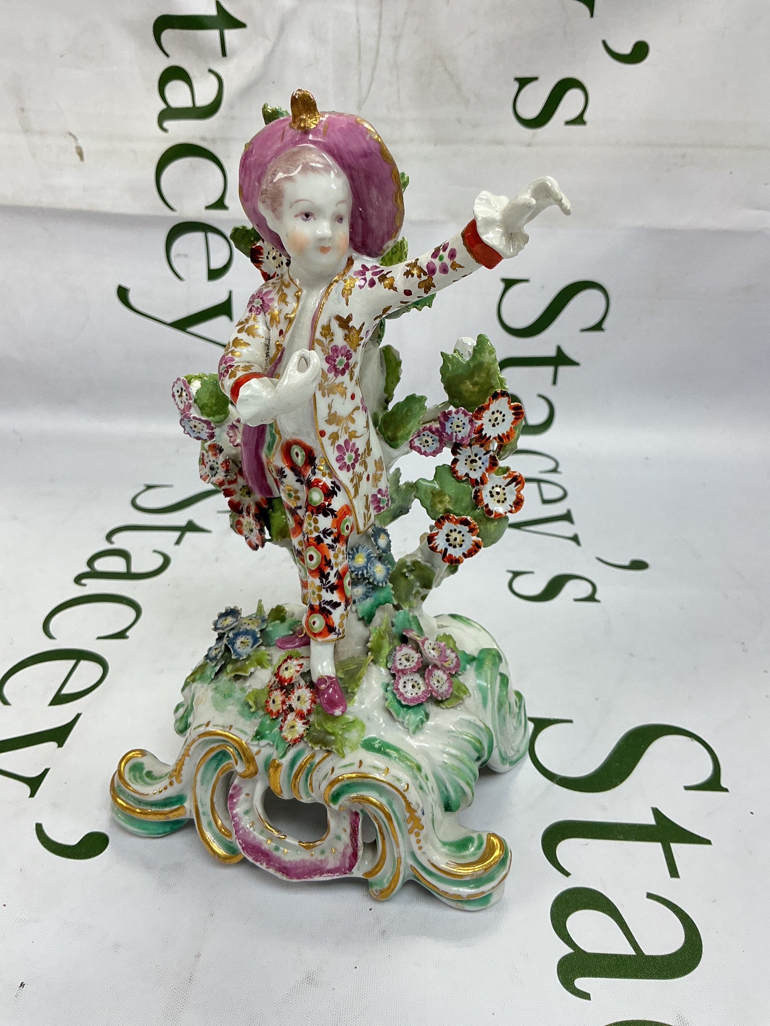 A circa 1765 Bow figure titled New Dancer. The standing figure wearing a broad brimmed upturned puce coloured hat with floral jacket and trousers and raised on a pierced scroll base applied with flowers and foliage. Chipped with some restoration. Approx height 20.5cm.