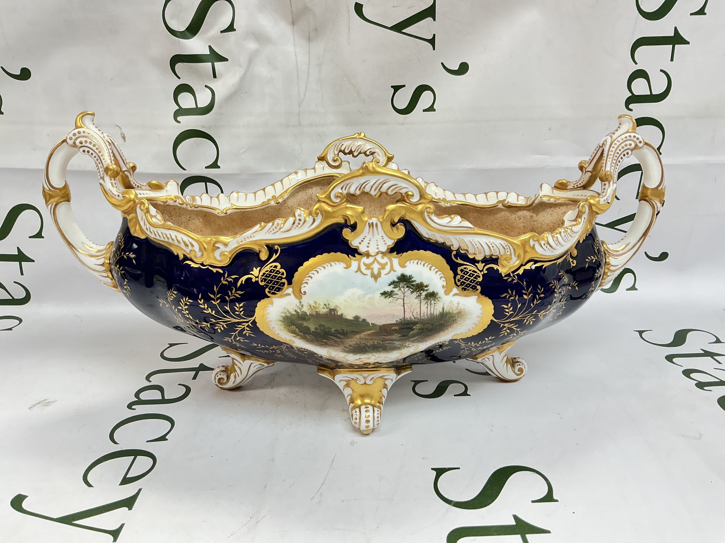 An early 20th century Coalport Hampton pattern two handled boat shaped table centrepiece, the central cartouche painted with a mountainous river scene, a flower spray to verso, resting on four scroll feet, approx 41cm wide, printed mark in green to base. Some restoration.