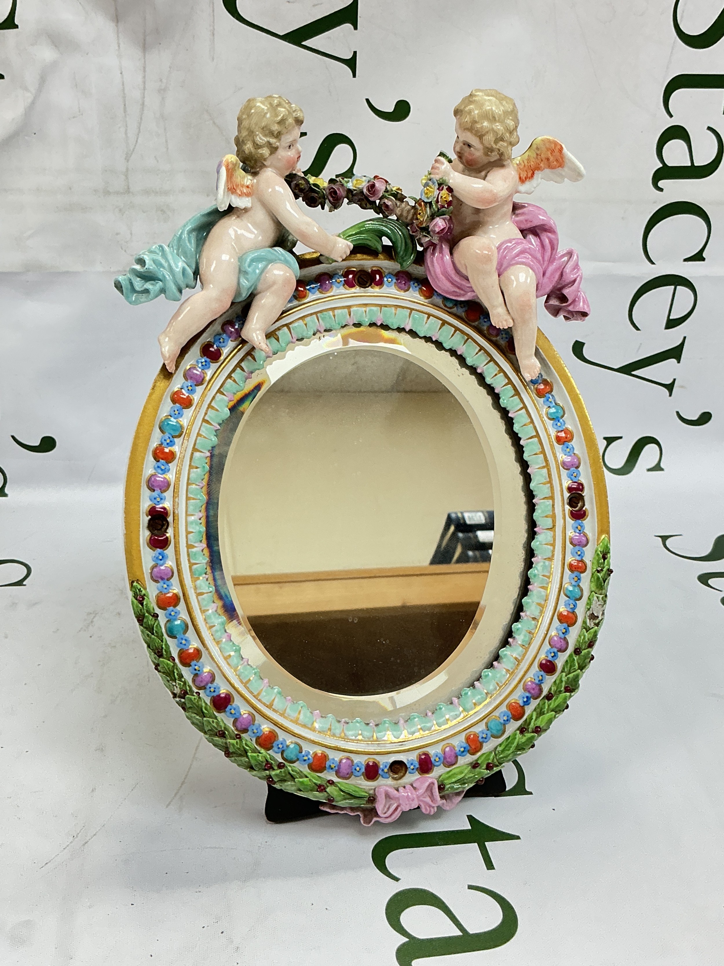 A Meissen table mirror of oval shape, surmounted by two cherubs holding a floral garland, the frame moulded and painted with stylised leaves, flowerheads and 'jewels'. Damaged. Approx height 25cm.