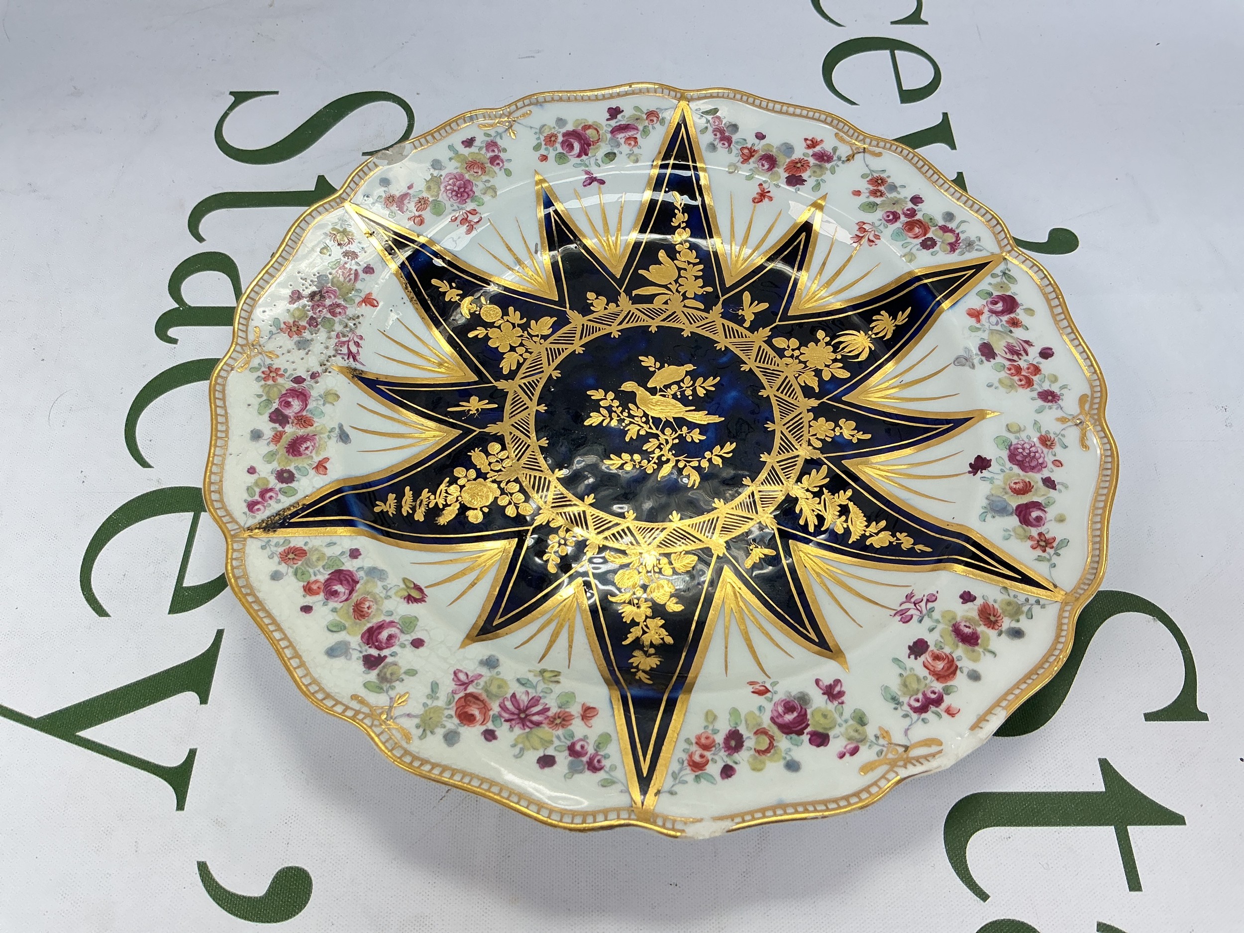 A mid 18th Century Chelsea plate with a large sunburst motif in Mazarin blue and gilt with birds and flowers, the rim with garlands of flowers, gold anchor mark to base, approx diameter 22cm. Chips and visible cracks to edge.