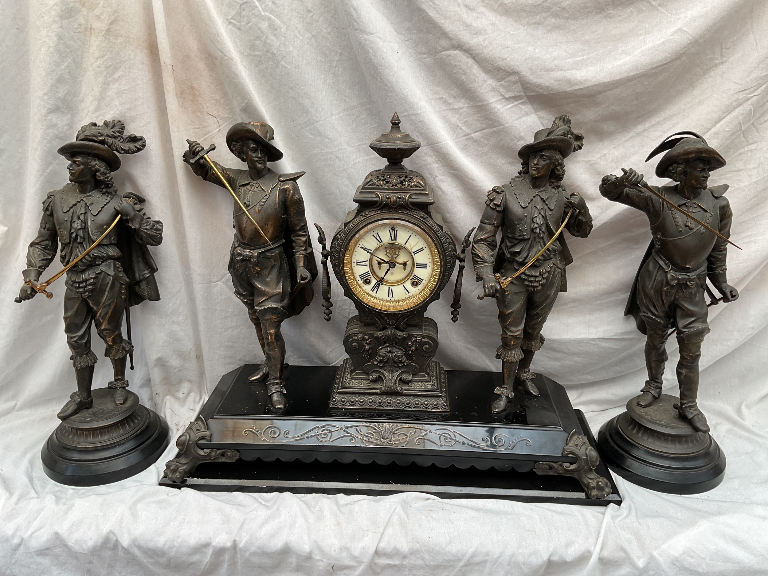 19/20th C Spelter clock garniture with Cavaliers and an Ansonia eight day movement striking on a gong, all on marble bases the central clock on a wooden plinth. Clock 54 cm high 63 cm wide,The side Cavaliers are 52 cm.