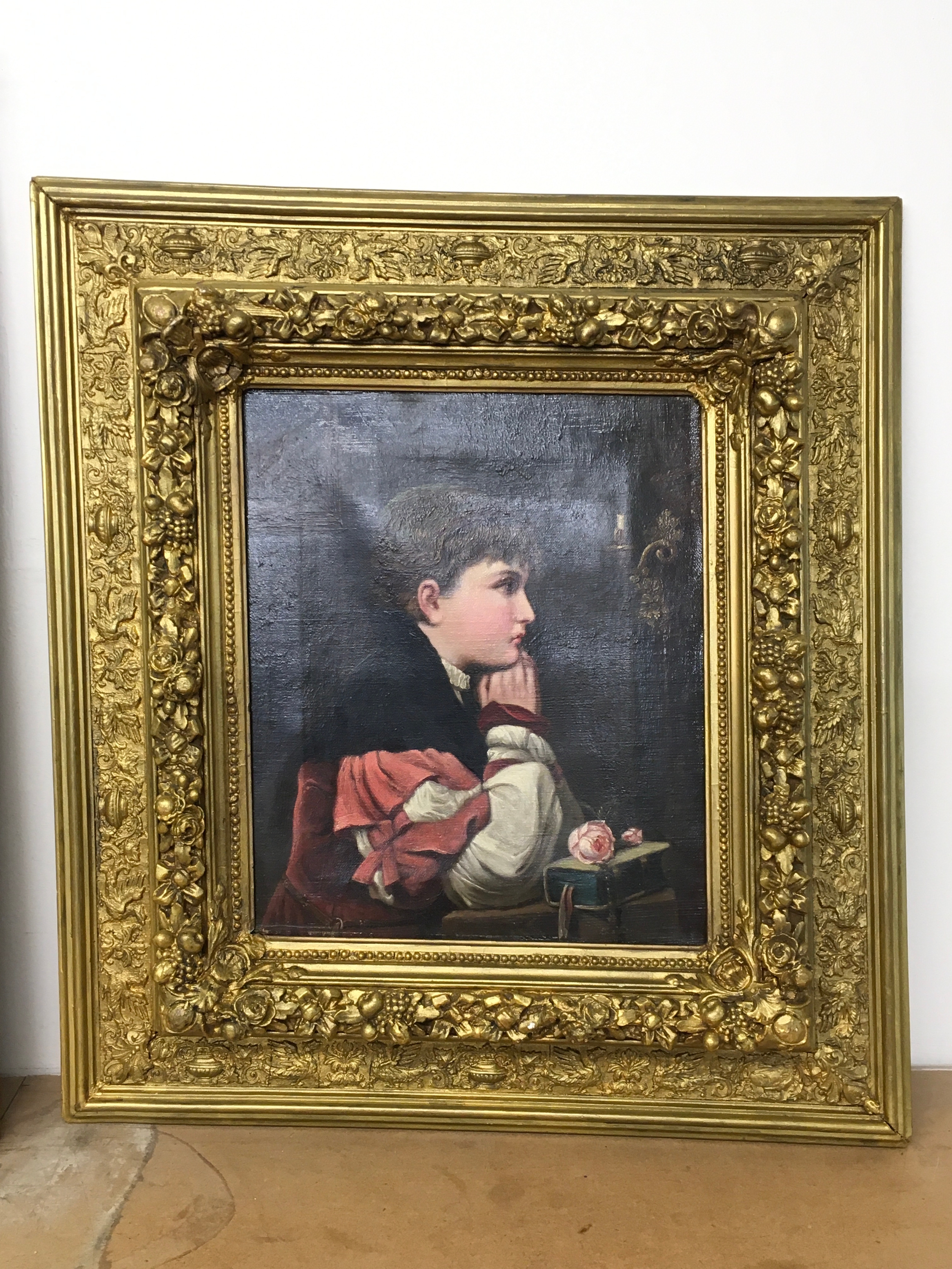 A 19th-century oil painting on canvas of a young boy resting by a book and a rose, housed in a heavily ornate, deep gilt-wood Baroque-style frame. Frame size 48x53cm picture 25x30cm