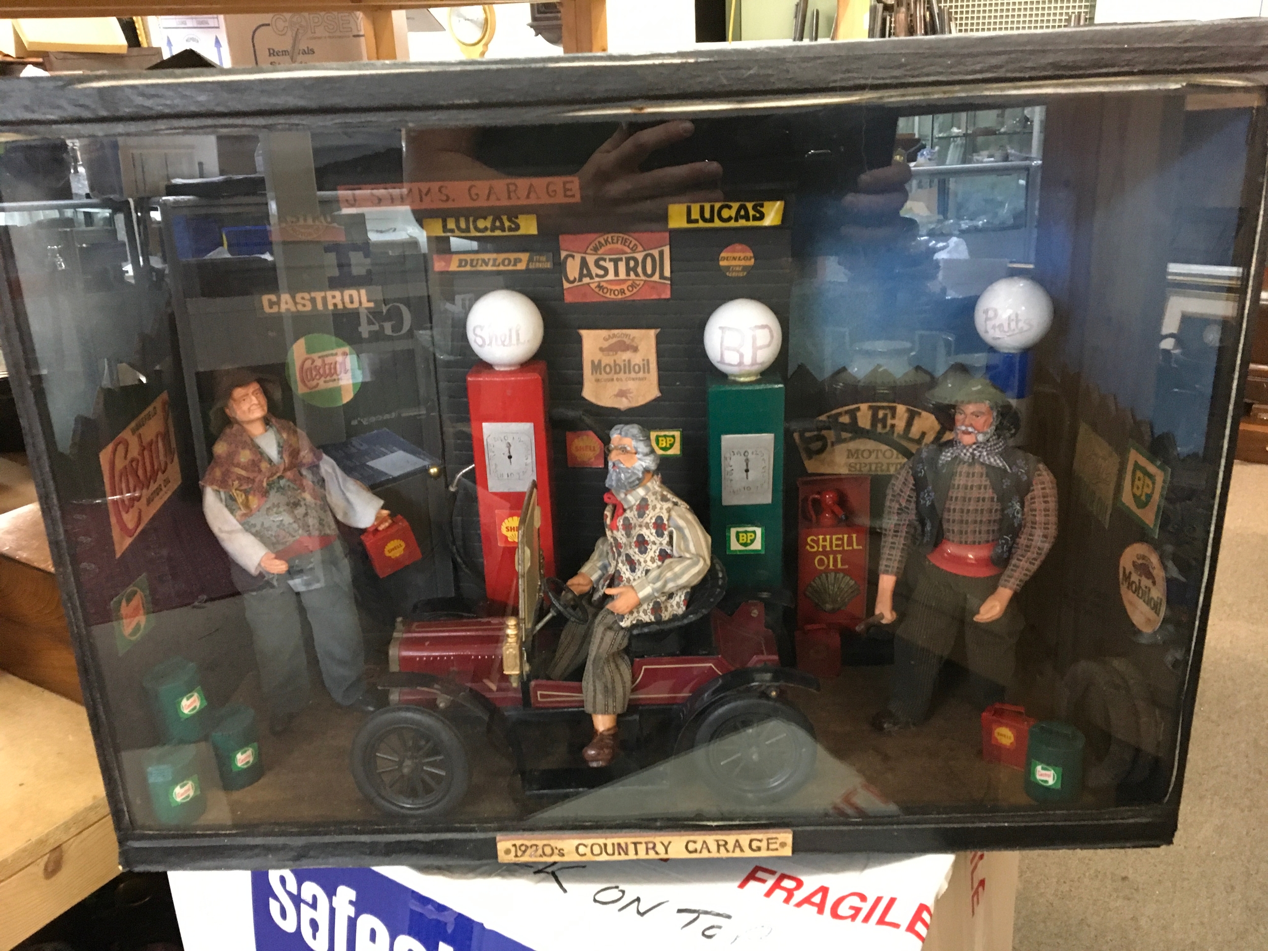 A diorama of a 1920s village garage with pumps, oil and petrol cans, signs etc. Three figures one serving an old car with petrol