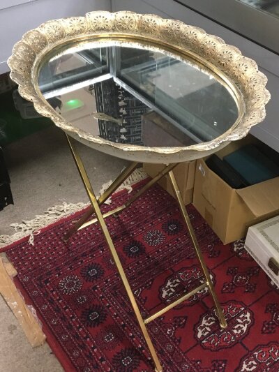 Moroccan brass tray table featuring a folding metal base and an ornate, oval-shaped mirror top. 71cm tall, 58cm wide. No reserve