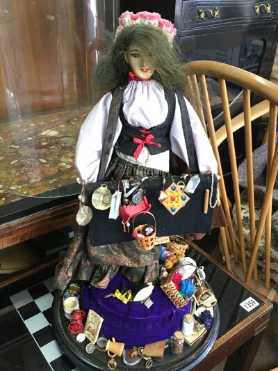 A Pedlar doll figure in a glass display dome, with an assortment of miniature trinkets. Dome is 67cm tall