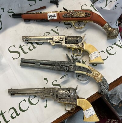 four non firing replica display guns. The flintlock at the top features a stained wood stock, a gold coloured cast metal filigree on the lock plate and a decorative flared metallic butt cap. The revolver second from the gun at the top has an octagonal barrel, a loading lever under the barrel, an engraved cylinder depicting a naval battle scene and a faux ivory grip. The second flintlock above the bottom one has two parallel antiqued-grey barrels featuring cast relief scrollwork, dual hammer and a highly decorative cream coloured faux-ivory grip carved with scrollwork and a gargoyle head on the pommel. The bottom revolver mimics a highly engraved luxury presentation model, featuring elaborate scrollwork across the silver-toned frame, a gold-plated trigger guard, and textured checkered faux ivory grips.